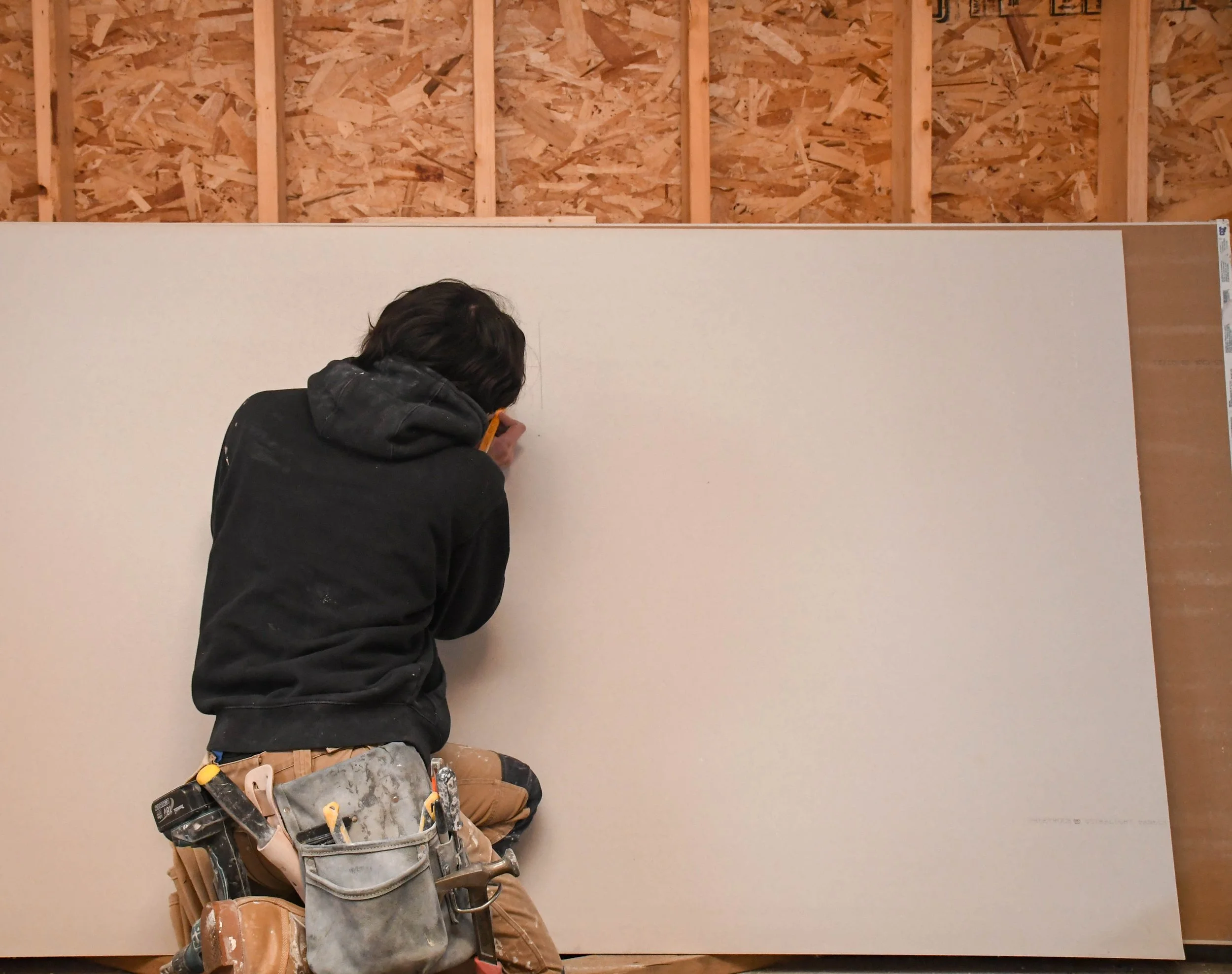 A construction worker wearing a tool belt, working on a large white drywall panel inside a building under construction.