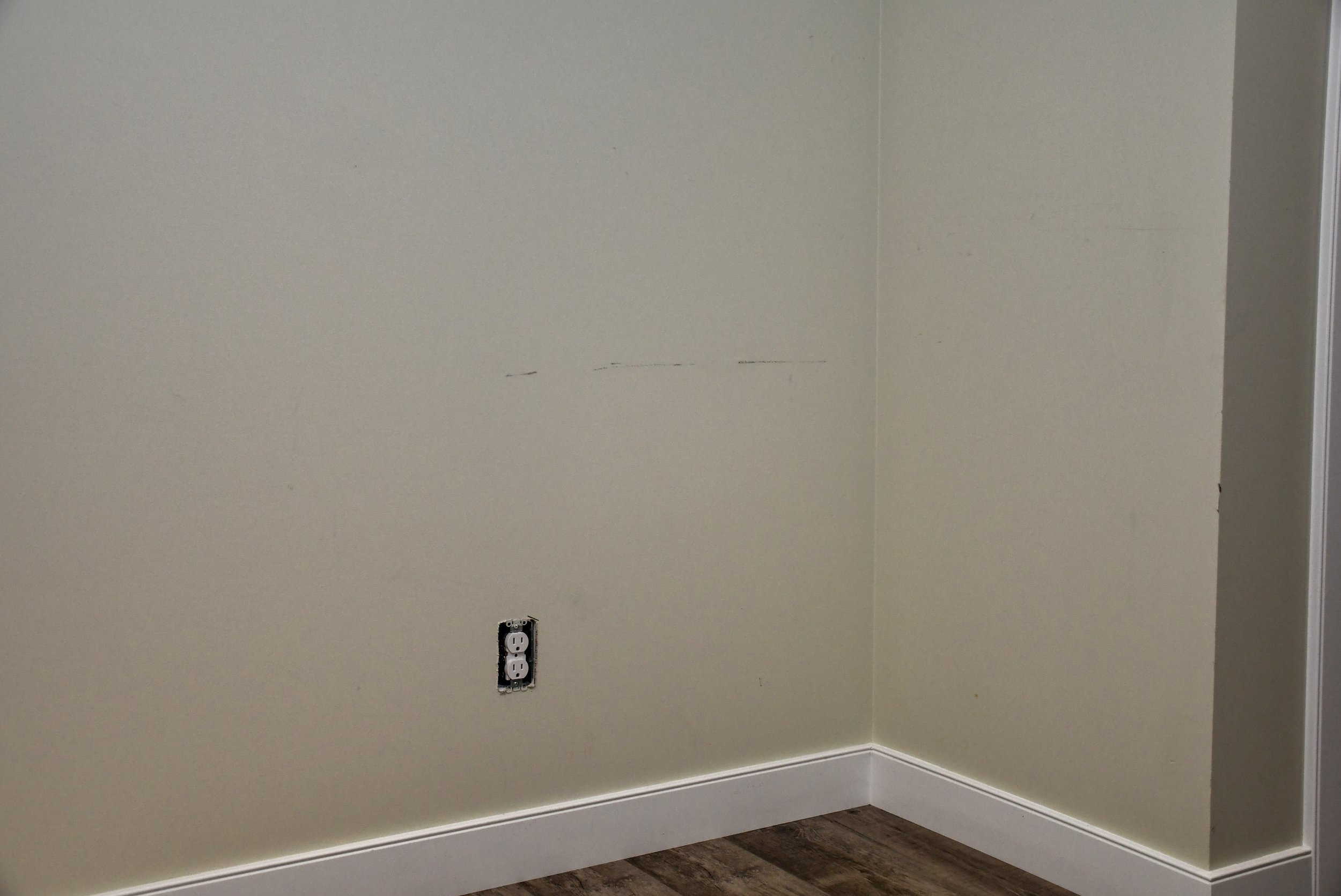 Empty room corner with a beige wall, a white baseboard, a damaged electrical outlet on the wall, and hardwood flooring.