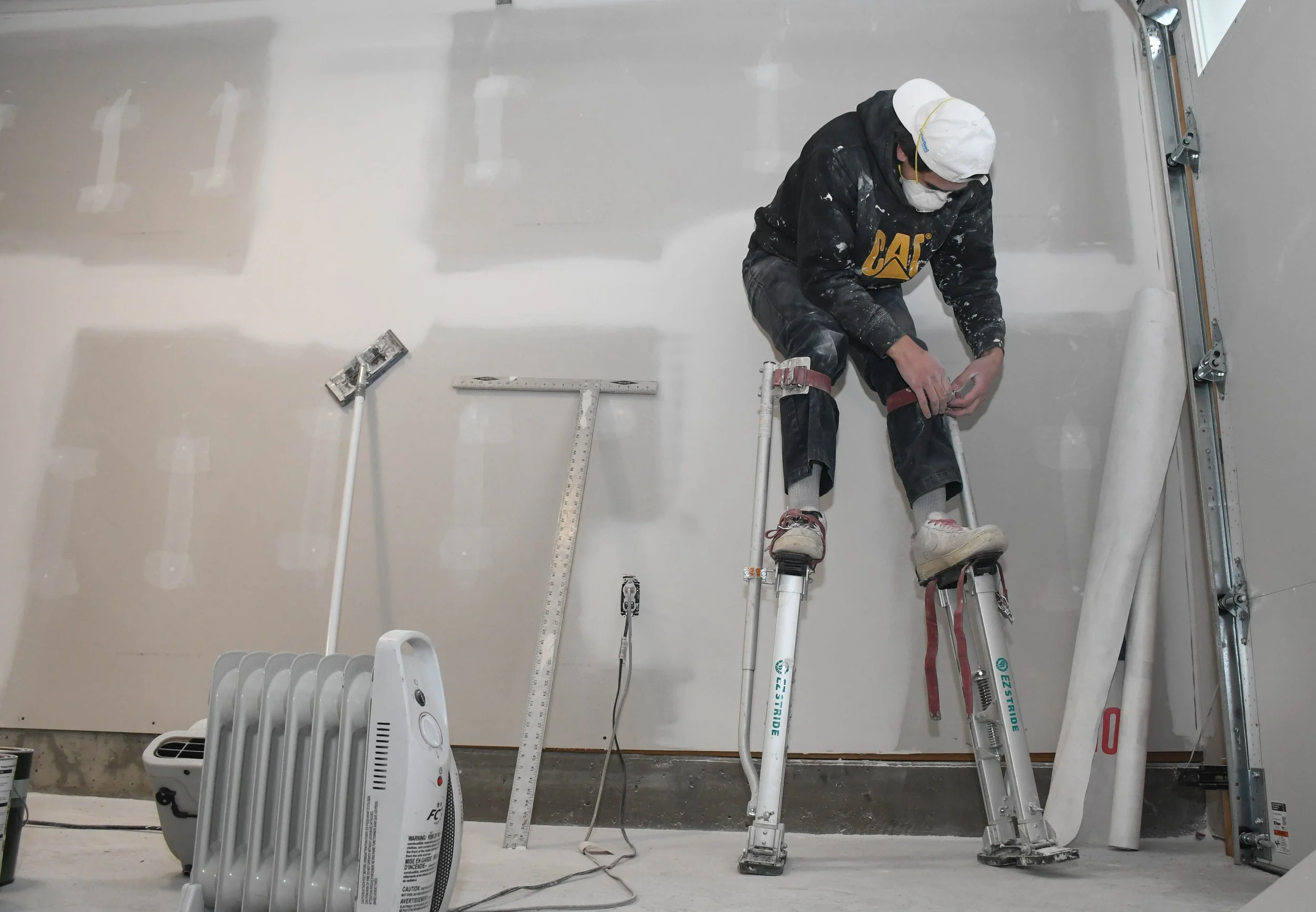 A worker with a white face mask and protective gear, sitting on stilts, installing drywall inside a construction site with tools and equipment around him.