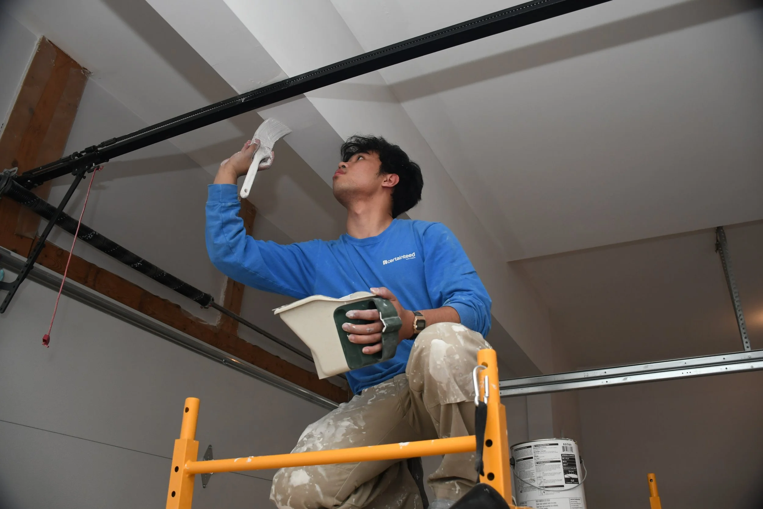 Man painting ceiling while sitting on a ladder with a paintbrush and paint roller container in hand.