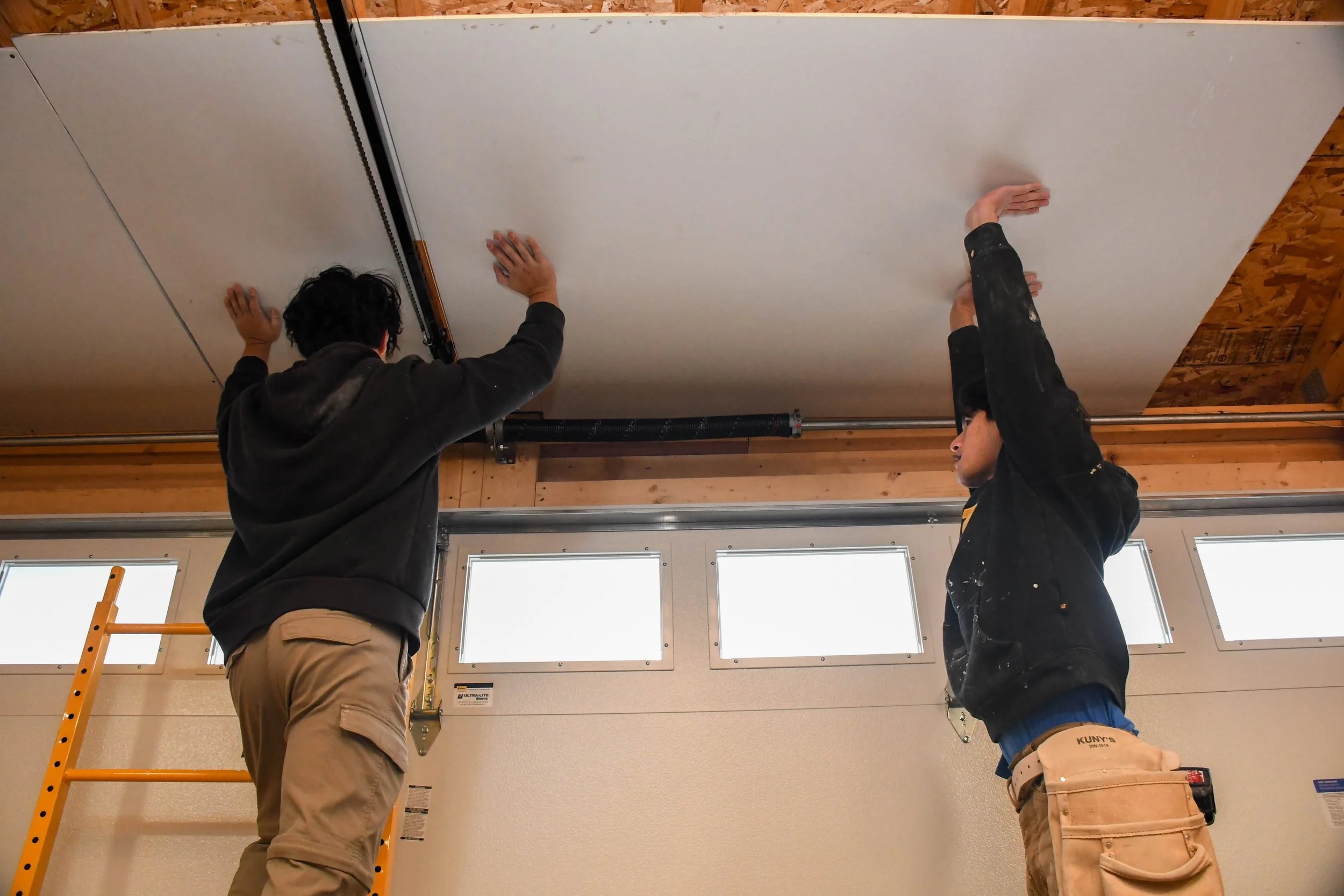 Two men working on installing or repairing a garage door, one on a ladder and the other on the ground, with both reaching up towards the ceiling.