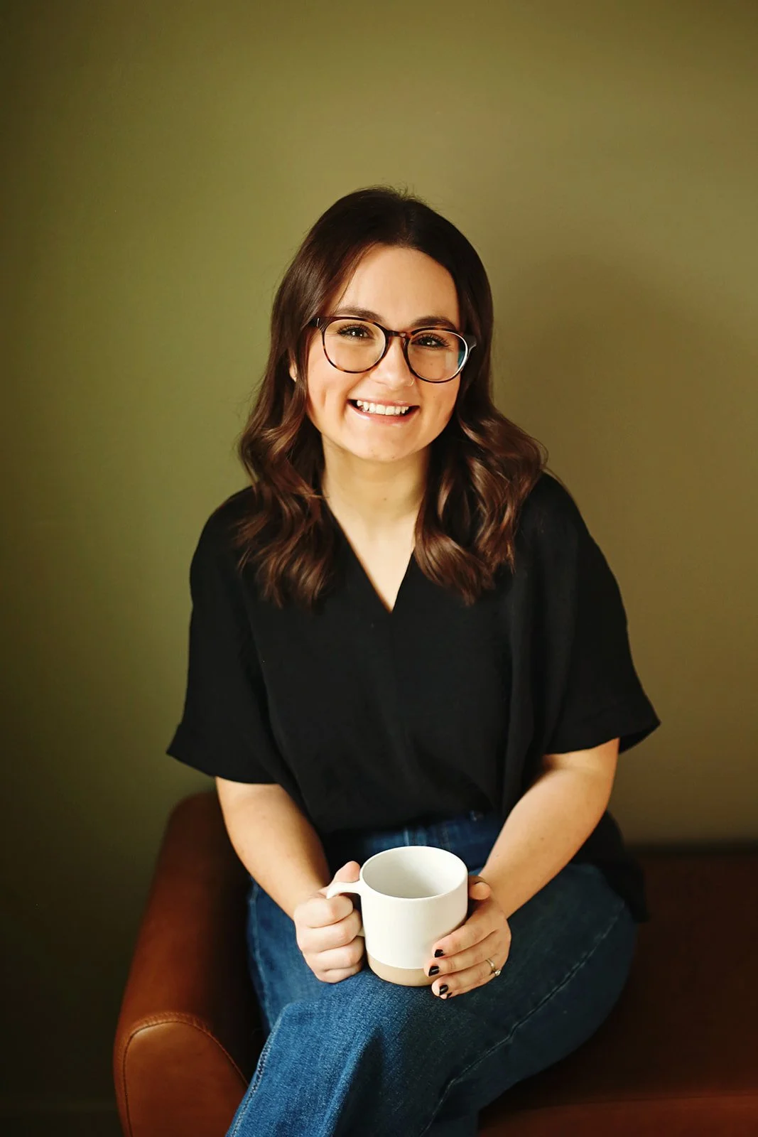 The speech therapist wearing glasses, holding a white mug, sitting on a brown chair, and wearing a black shirt and jeans.