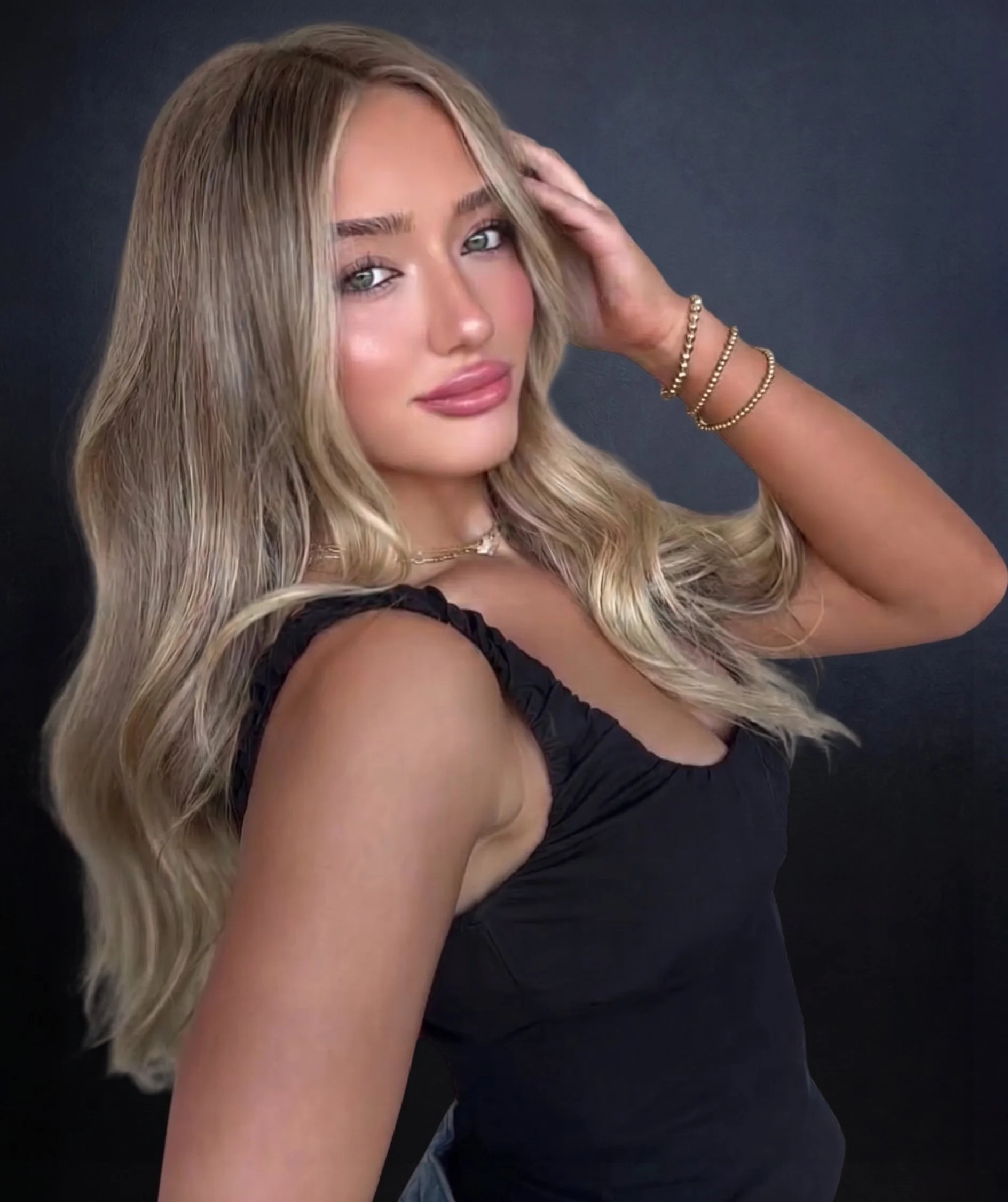 A woman with long, blonde hair styled in soft waves, posing against a plain dark background. She is wearing a black sleeveless top and gold jewelry, including bracelets and a necklace. She has light makeup, with pink lipstick and subtle eye makeup, and is looking directly at the camera with a slight smile, with her left hand resting on her head.
