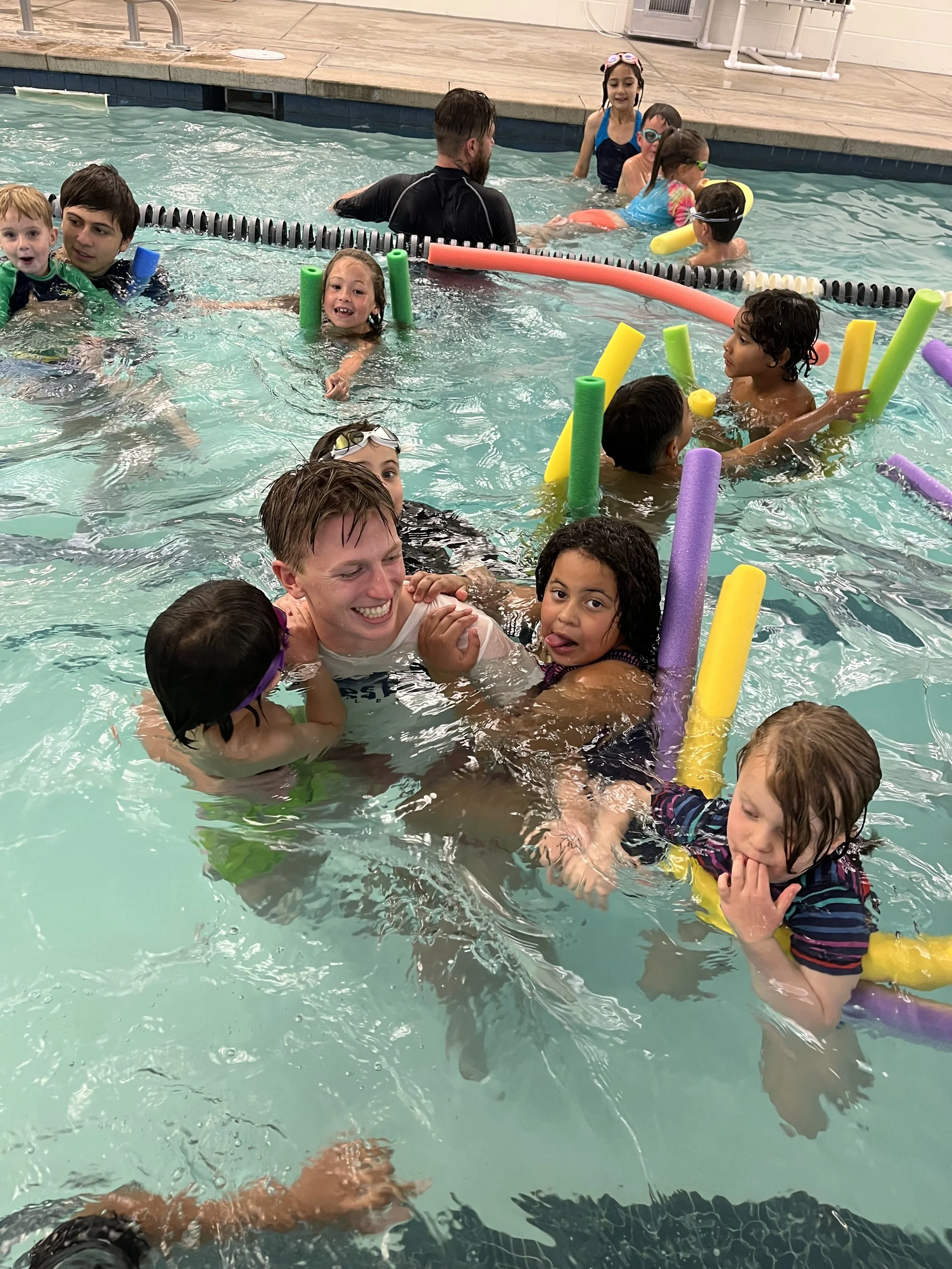Children and adults enjoying a swimming lesson in a pool with pool noodles and flotation devices.