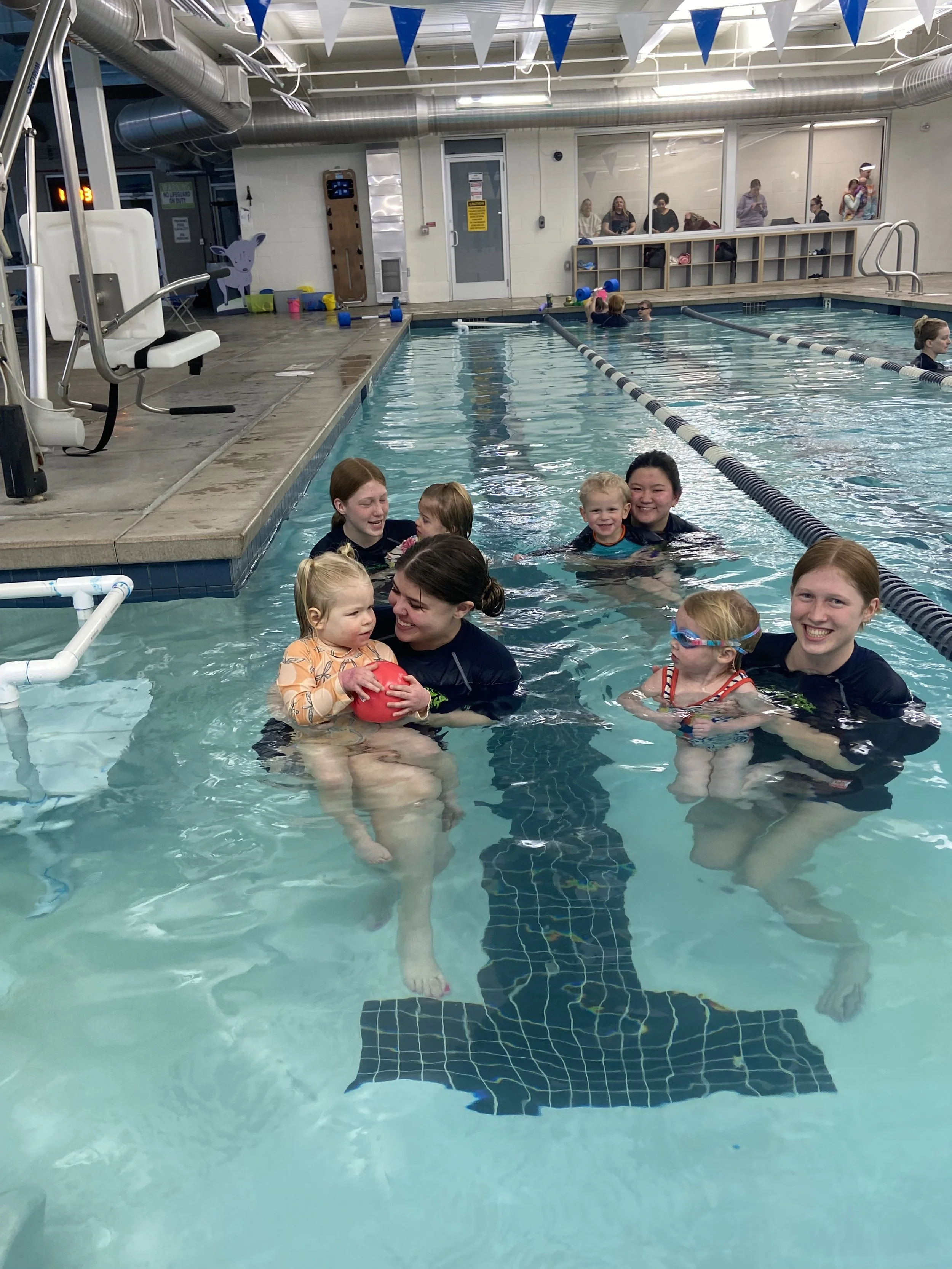 Group of children and adults enjoying a swimming lesson or activity in an indoor pool, with some paired with instructors and others swimming independently, surrounded by pool equipment and spectators in the background.