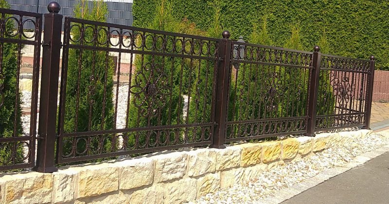 Una cerca de hierro forjado con diseños decorativos montada sobre un muro de piedra, rodeando un jardín con arbustos verdes.