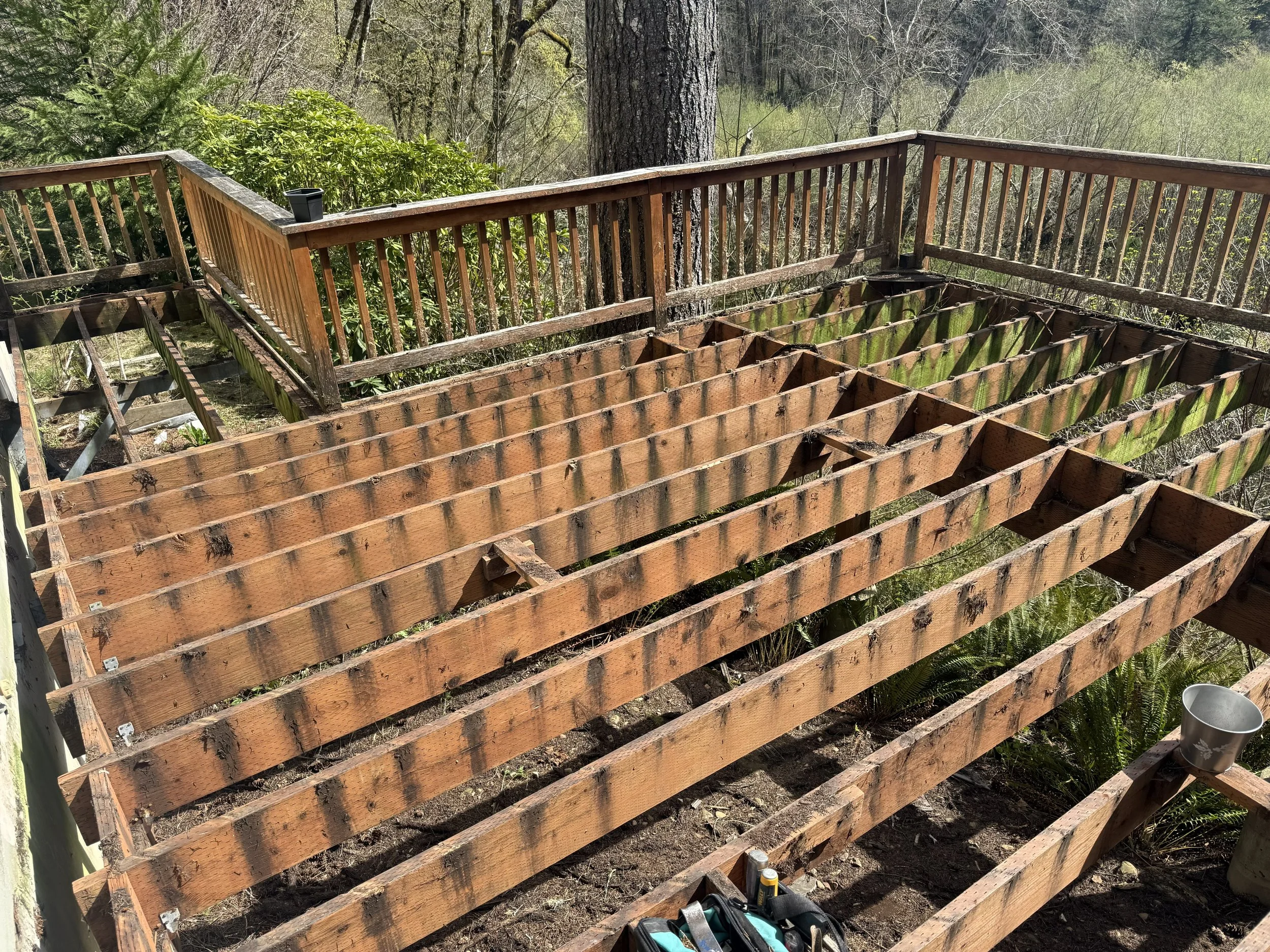 A wooden deck under construction in a wooded area, with some of the floor framing in place and tools nearby.