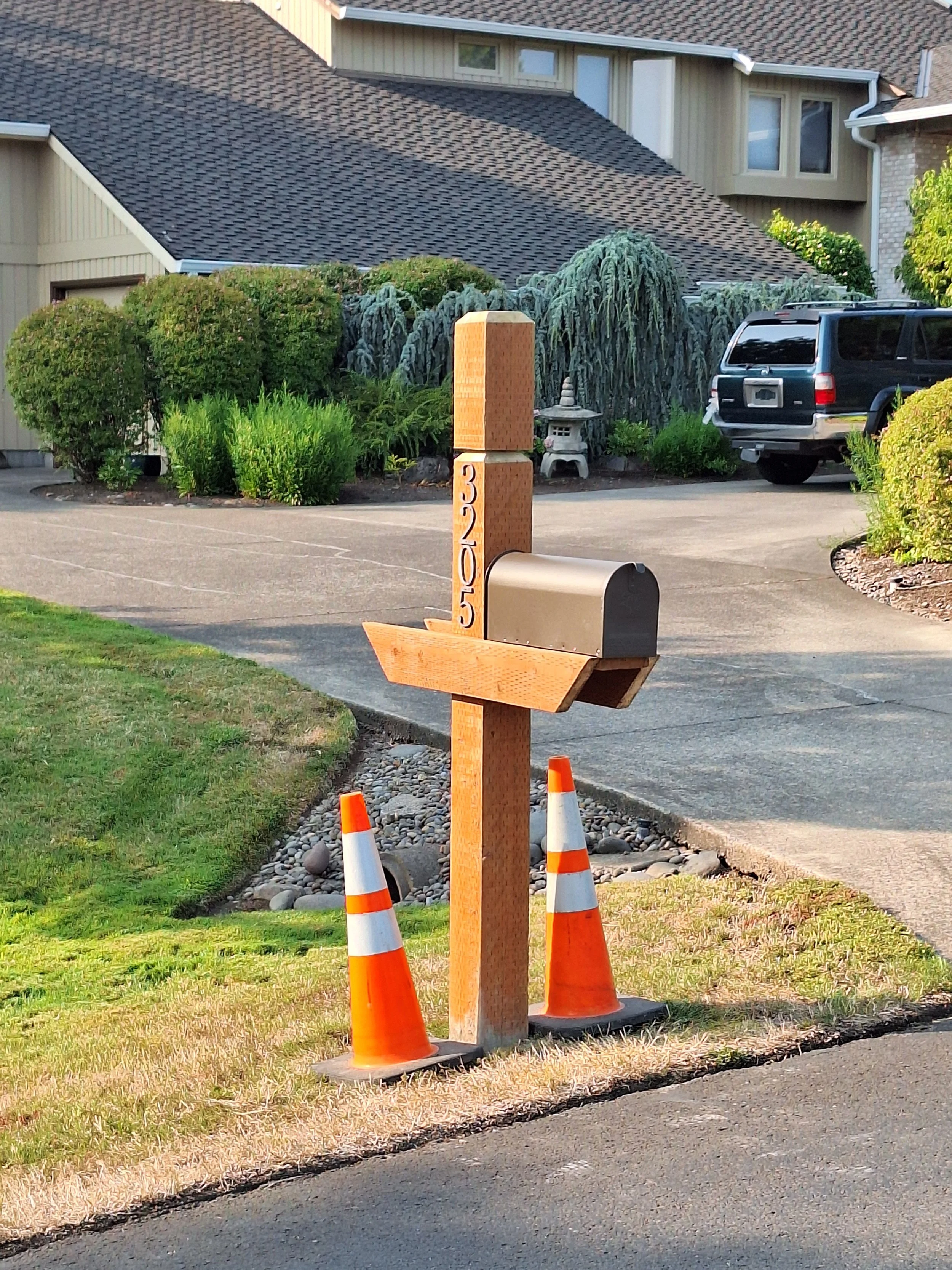 A suburban mailbox on a wooden post with writing on it, next to two orange and white traffic cones, in front of a house with greenery and a parked vehicle in the driveway.