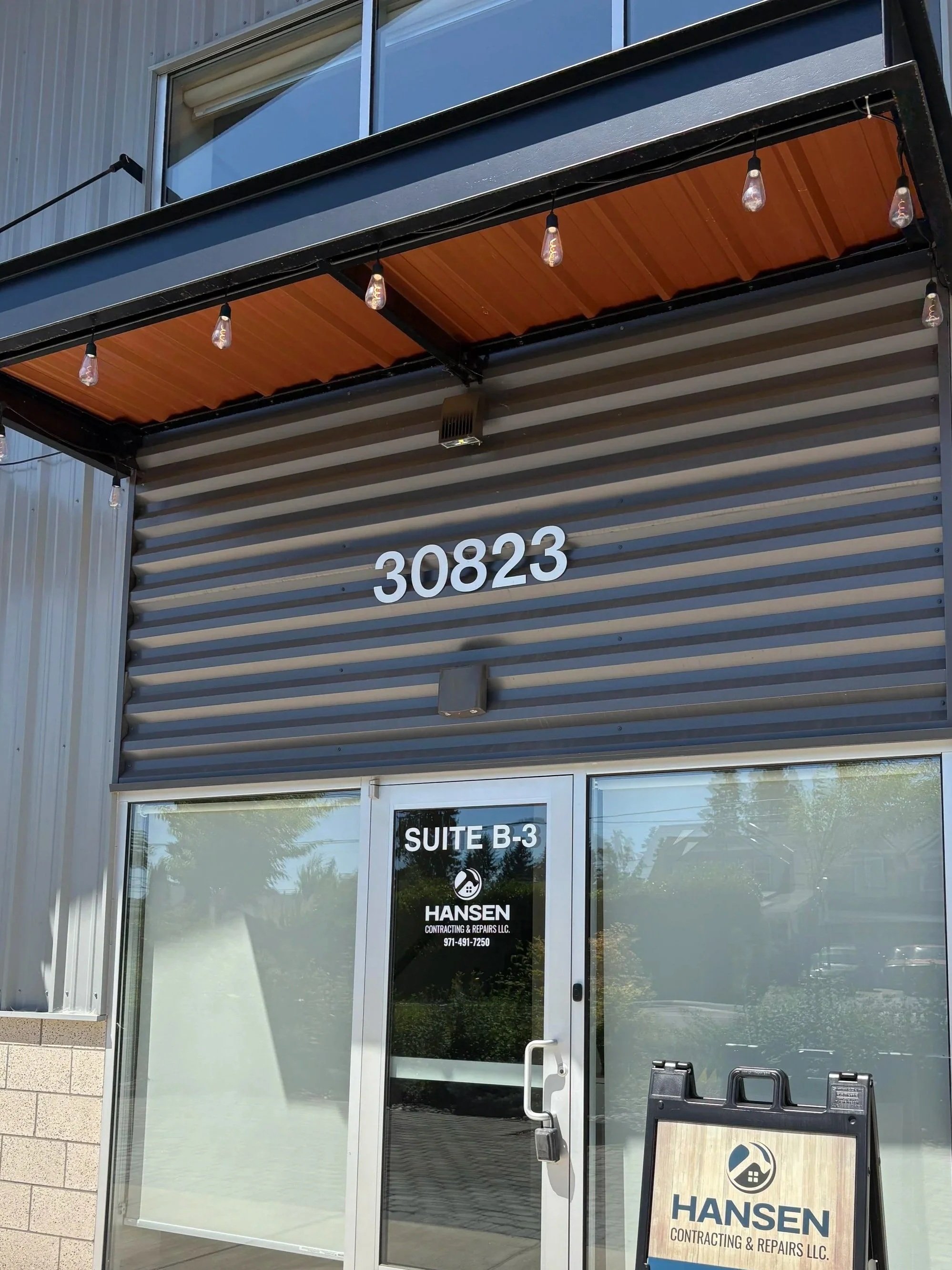 Photo of a building entrance with a sliding glass door, displaying the address 30823 and a black sign for Hansen Contracting & Repairs LLC, including a logo of a house and a wrench. Above, there is a balcony with string lights, and an air vent is visible on the exterior wall.