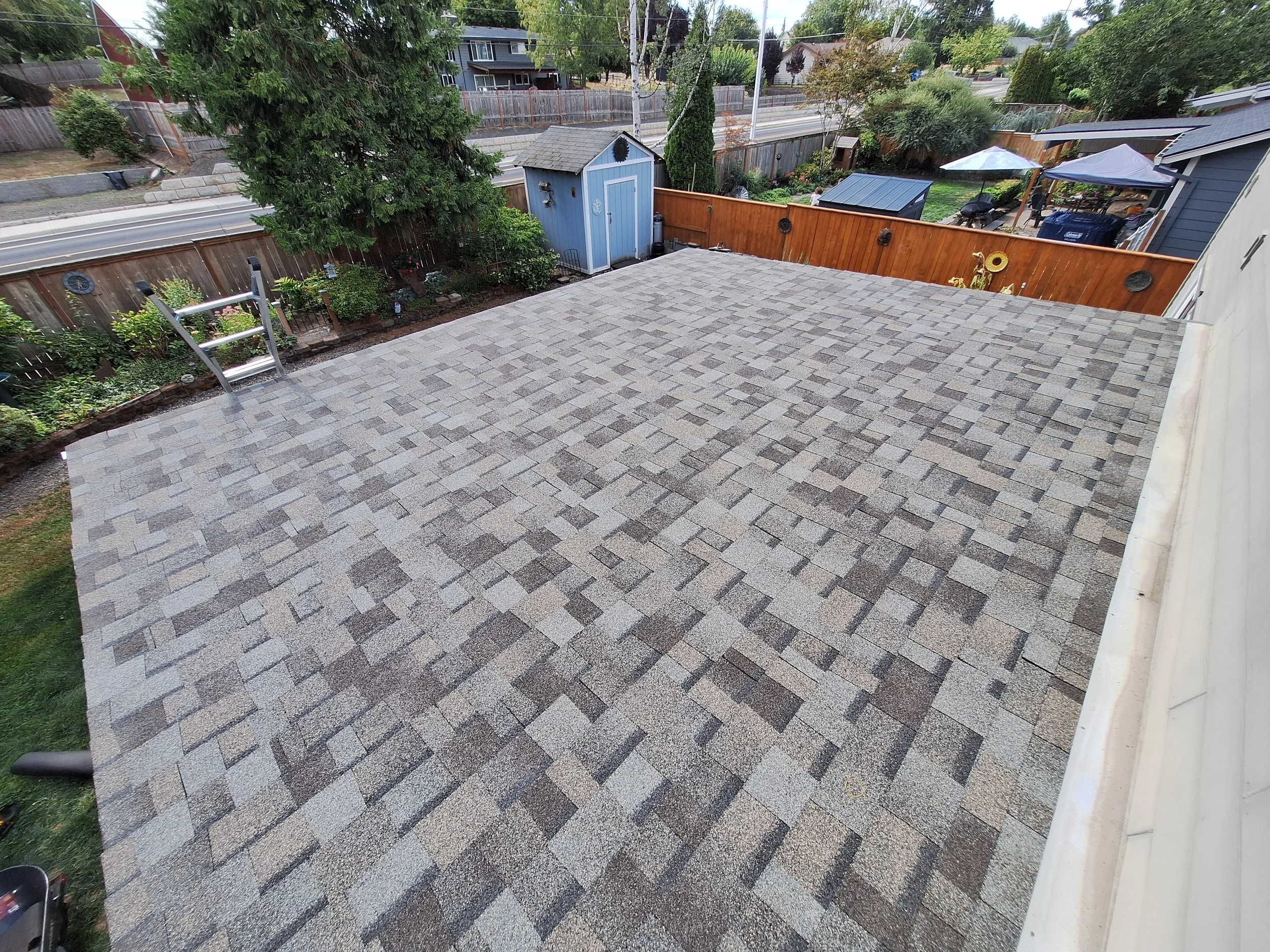 View of a residential backyard with a newly installed gray shingled roof and a children's ladder, surrounded by a wooden fence, garden beds, trees, and neighboring houses.