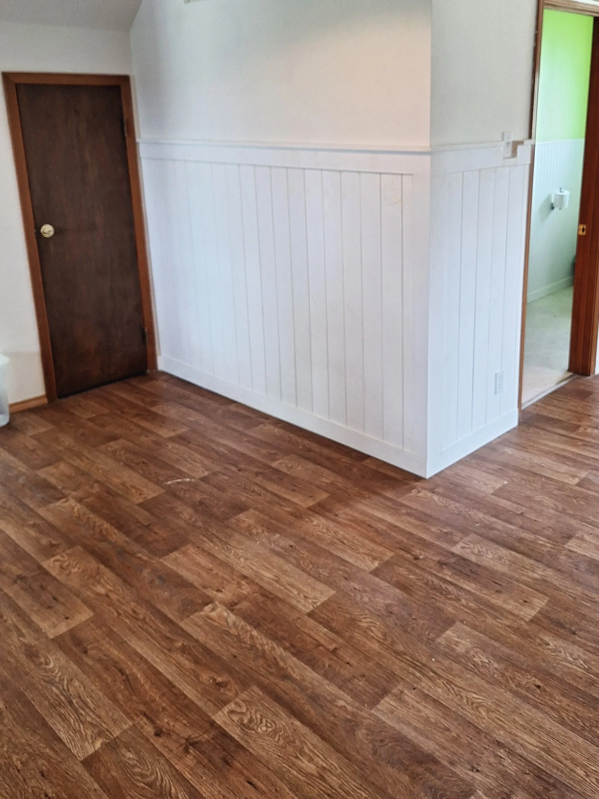 Interior view of a room with hardwood flooring, white walls with beadboard paneling, and two wooden doors, one dark brown door on the left side and one partly visible door on the right side leading to another room with light green walls.
