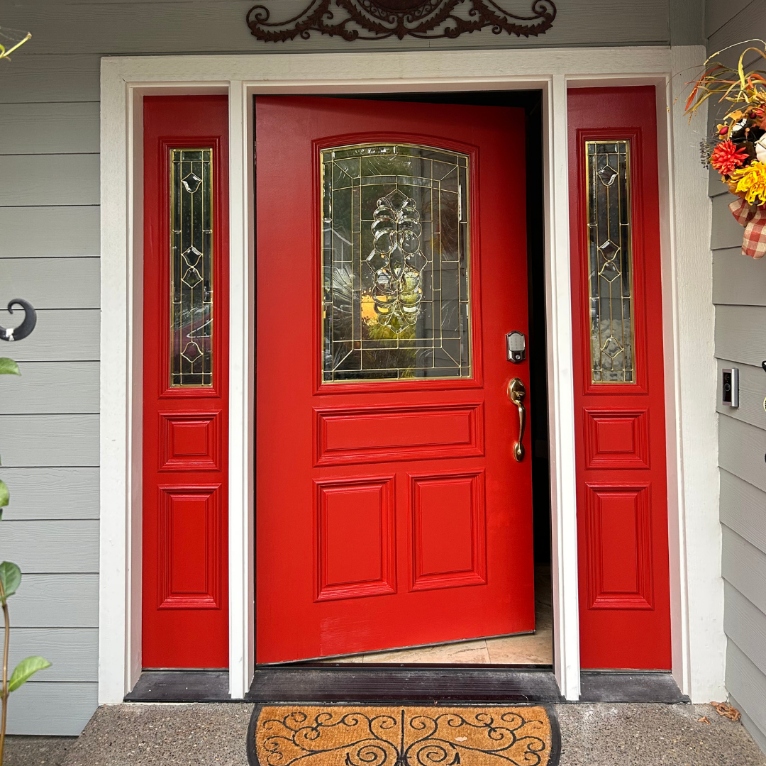 Red front door with decorative glass panels, surrounded by light gray siding and white trim, with a floral wreath on the right side.