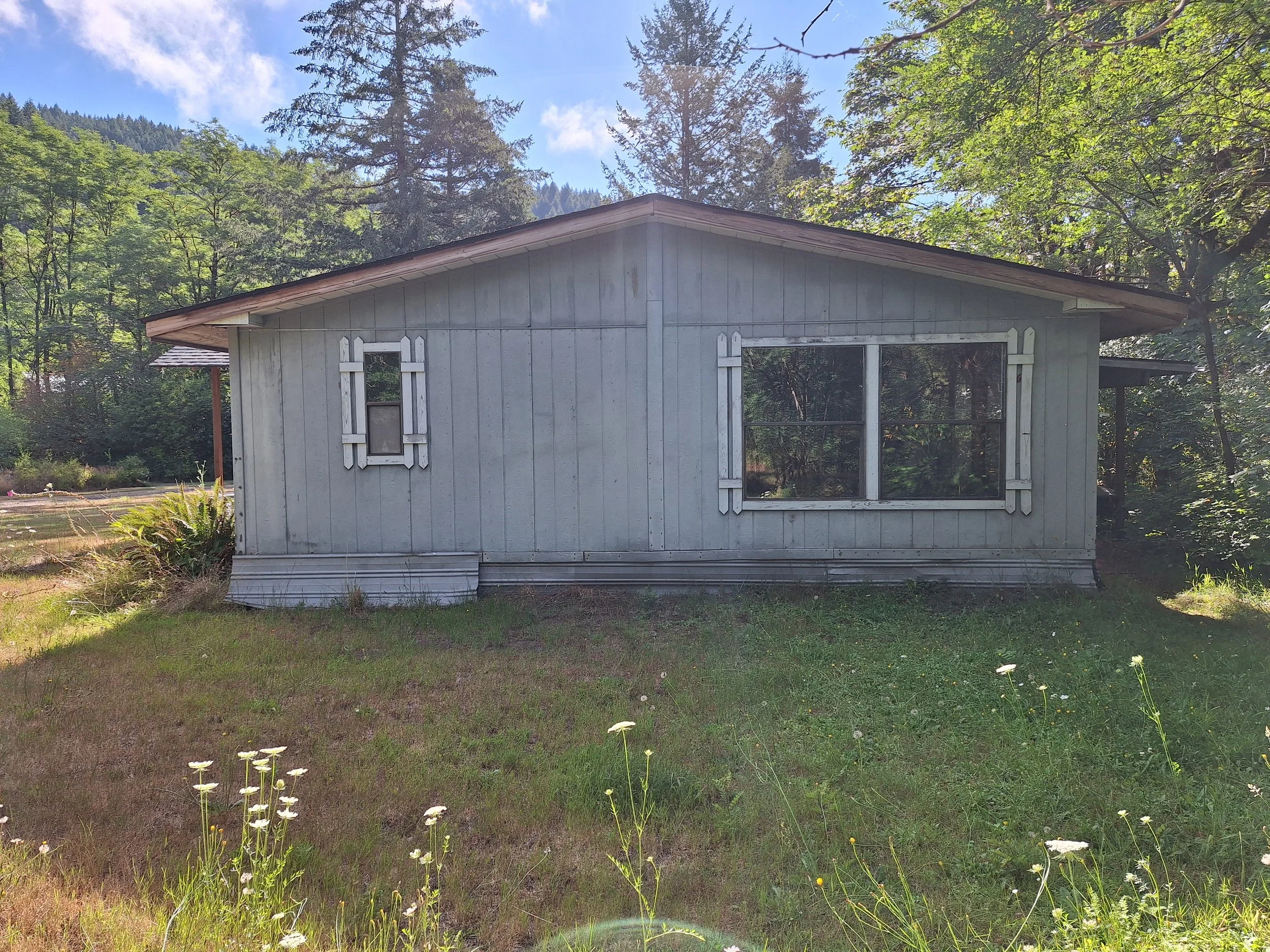 A small house with a gray exterior, decorative white window shutters, and a gable roof, set in a lush green landscape with tall trees and a bright blue sky.