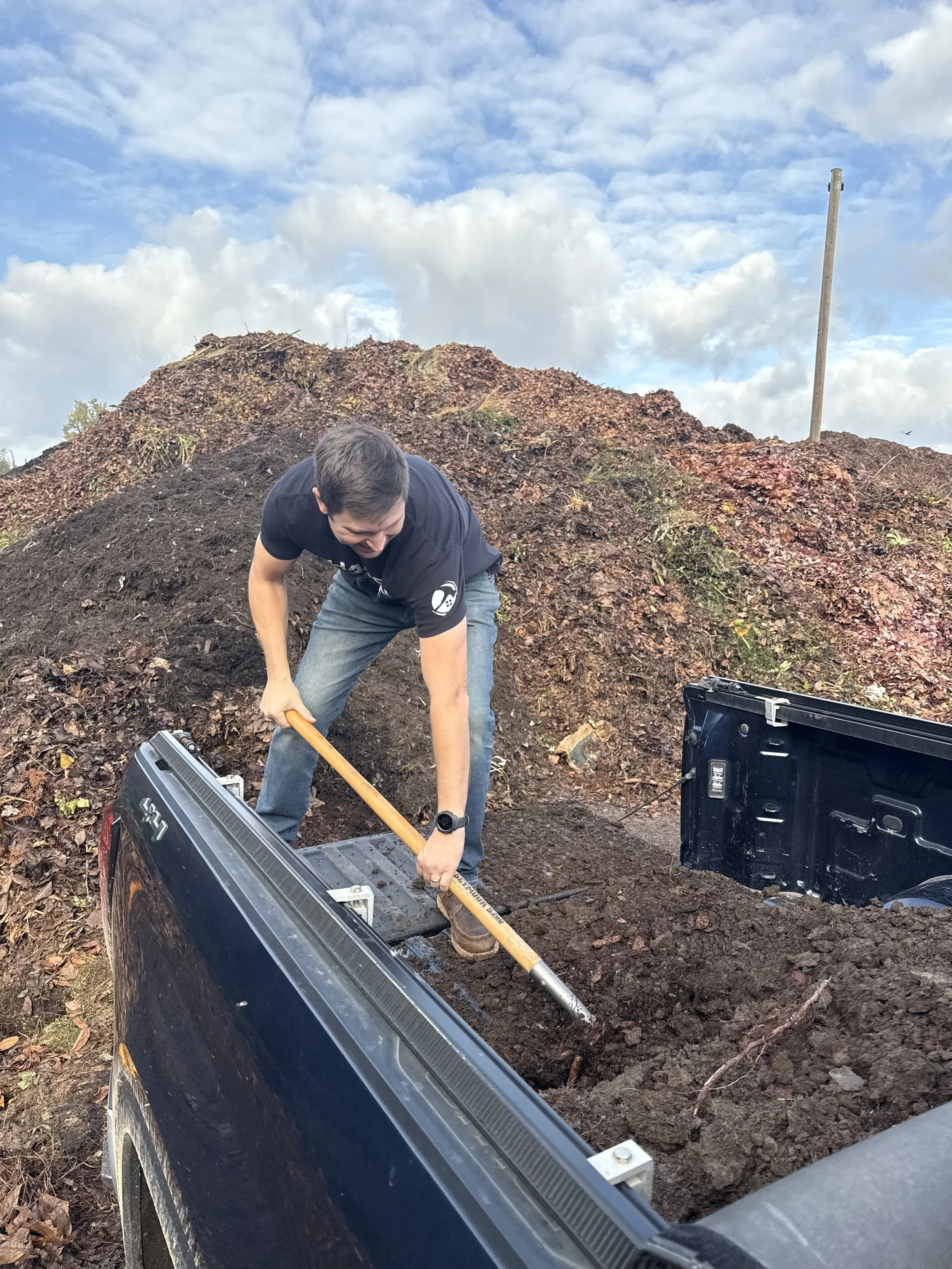 A man is digging in a hole with a shovel in a dirt area near a pickup truck. There is a large pile of dirt and leaves behind him, and the sky is partly cloudy.