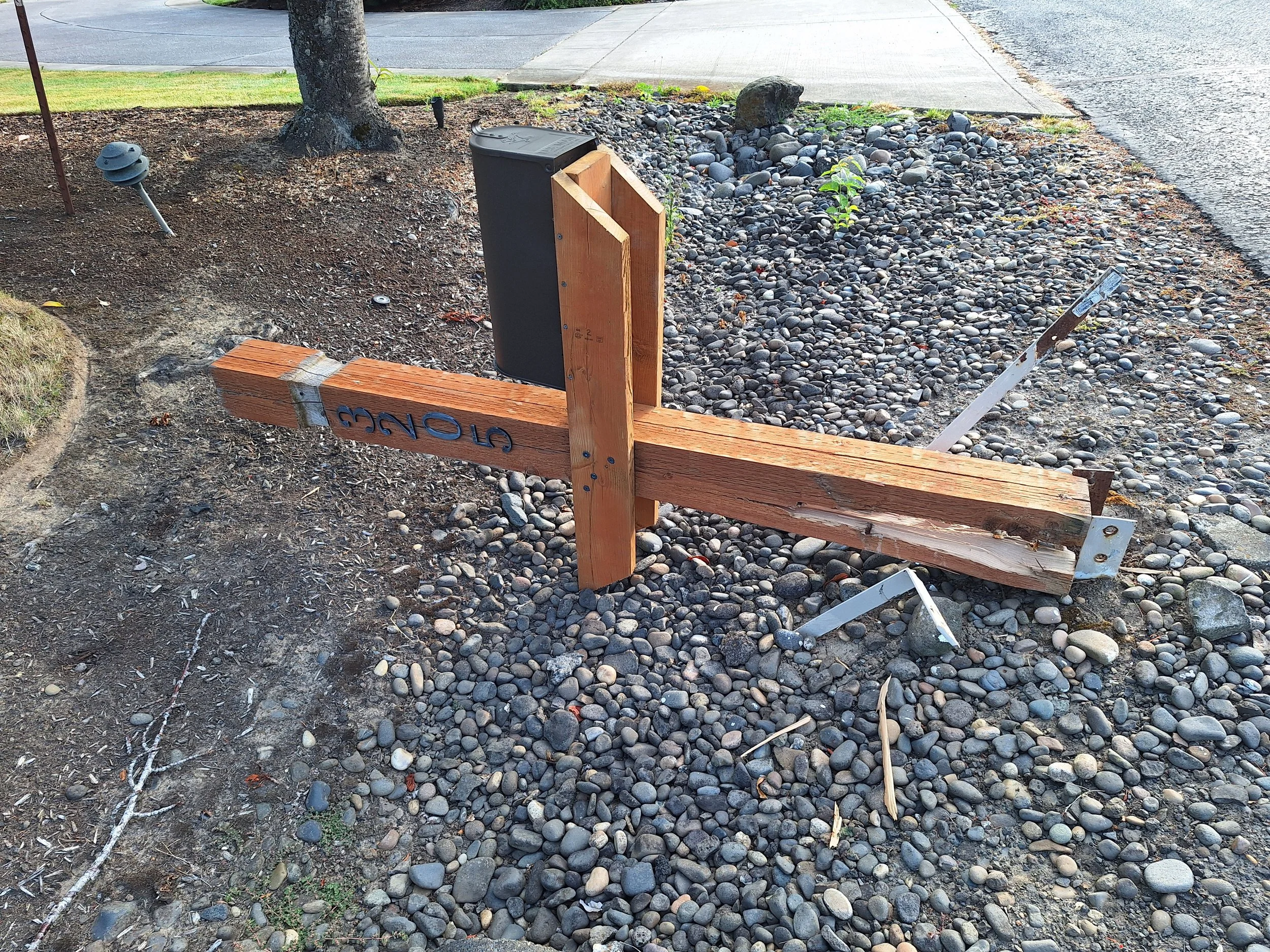 A street sign that has fallen over on a bed of small rocks and dirt, with a wooden post and metal brackets, next to a sidewalk and a tree. The sign is upside-down, with the word 'GONE' visible.