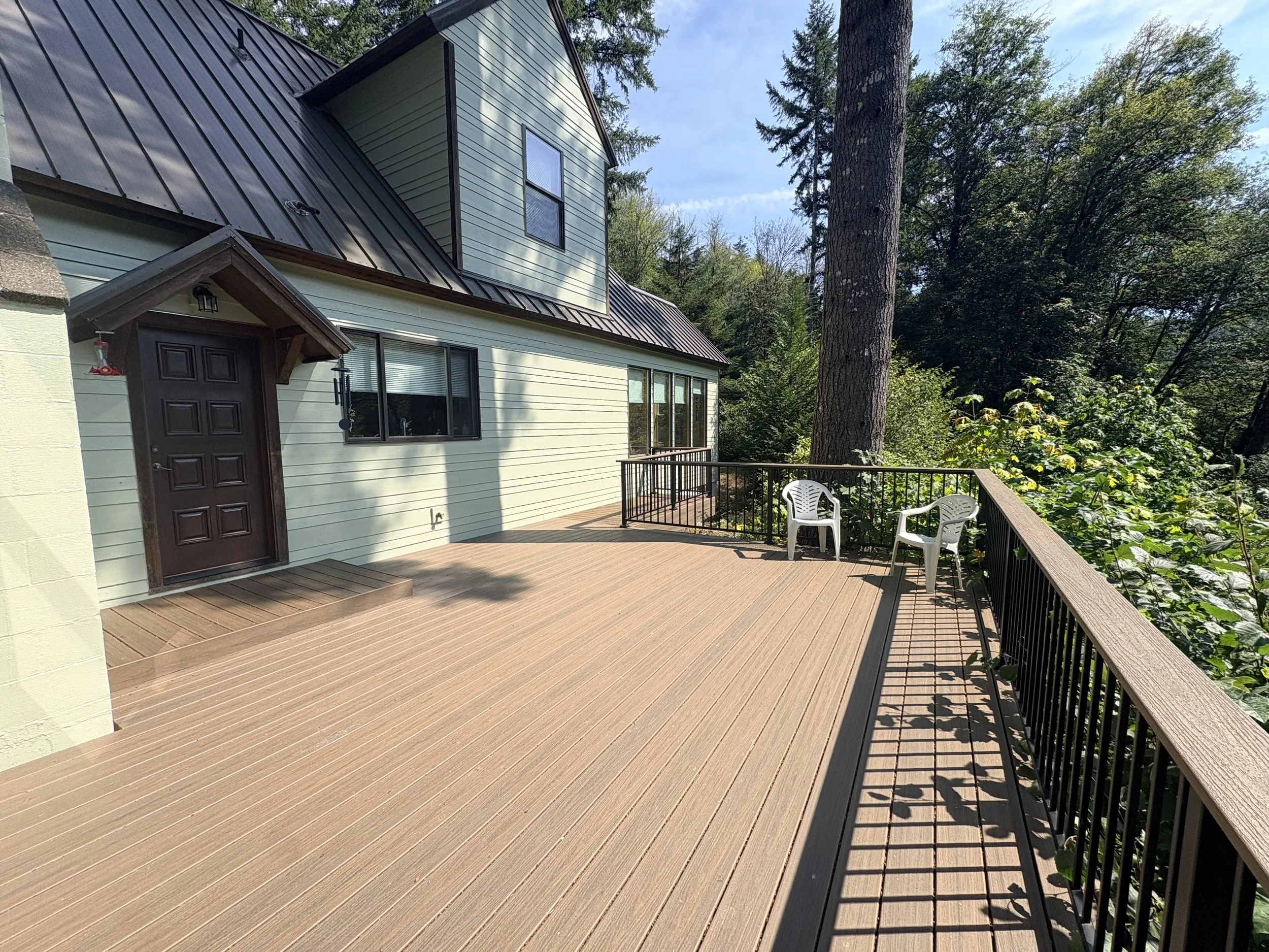 A house with a metal roof, green siding, and a deck with outdoor chairs surrounded by trees.