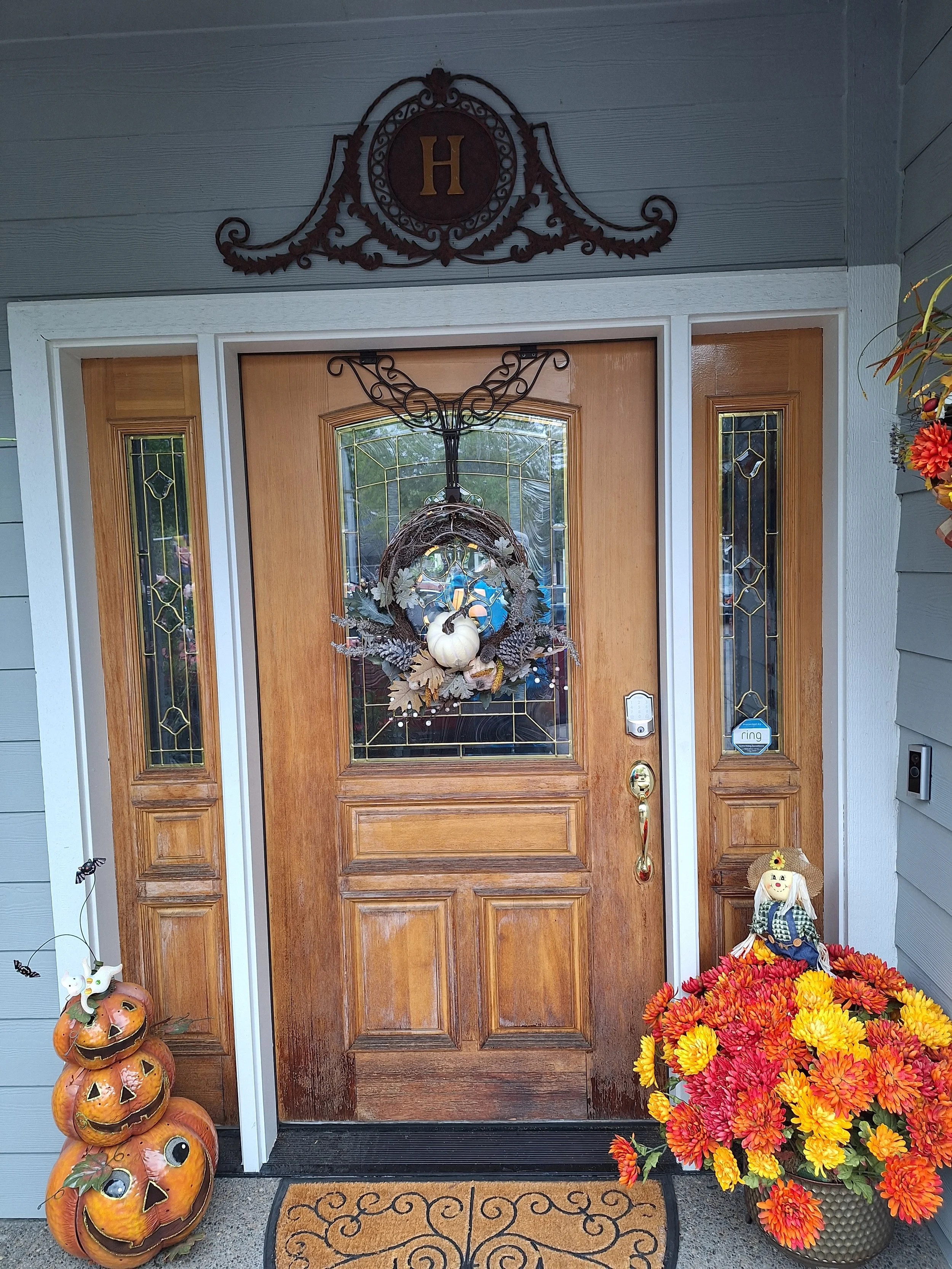 Decorated wooden front door with a Halloween and autumn theme, featuring a pumpkin wreath with fall leaves and flowers, a stack of three carved pumpkins on the porch, and a scarecrow doll among orange and yellow flowers.