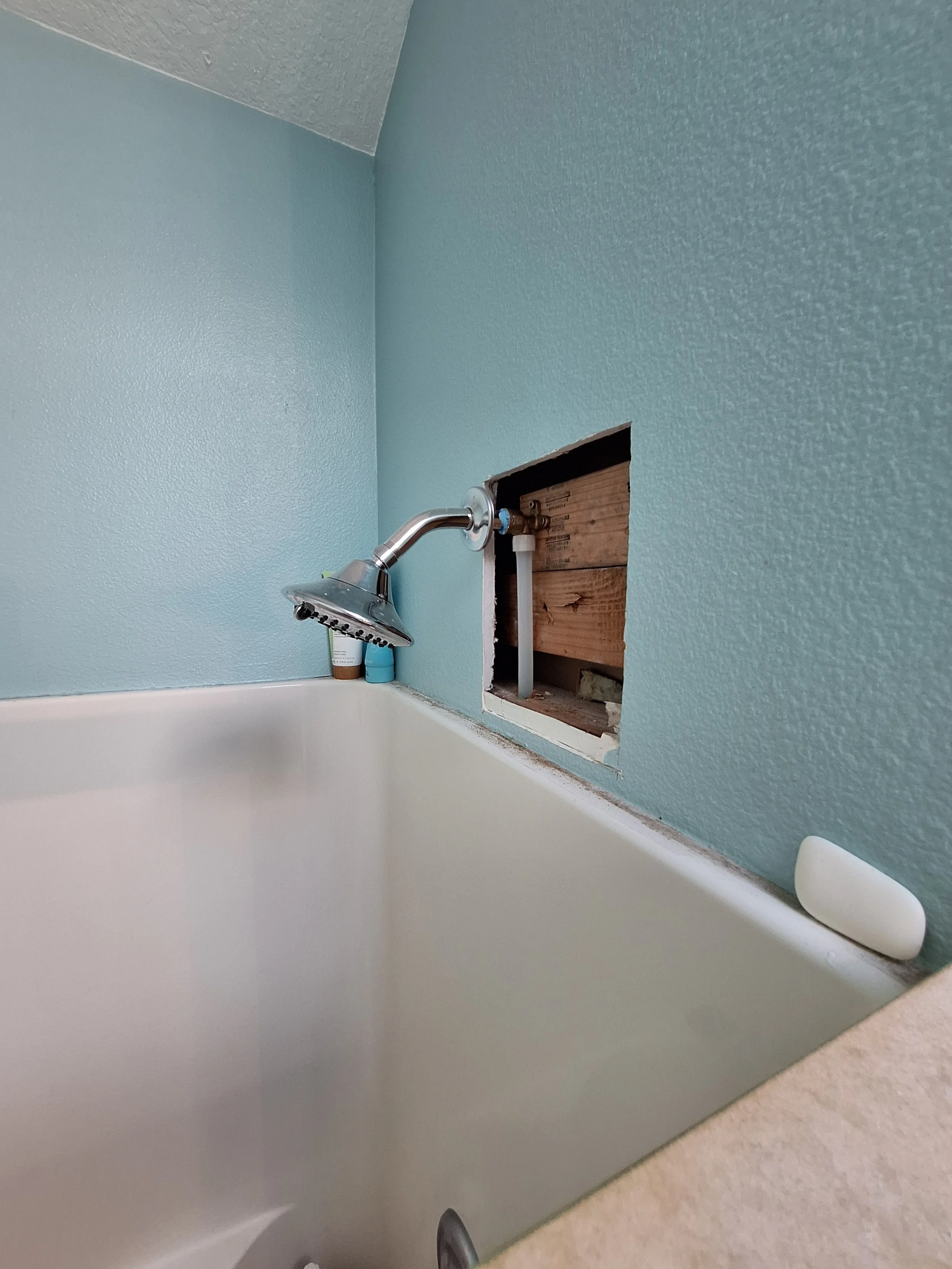 A bathroom with a bathtub and a light blue textured wall. There is an unfinished square opening in the wall, revealing wood and piping. A showerhead is attached to the wall near the open square, and two bottles are placed on the bathtub ledge.