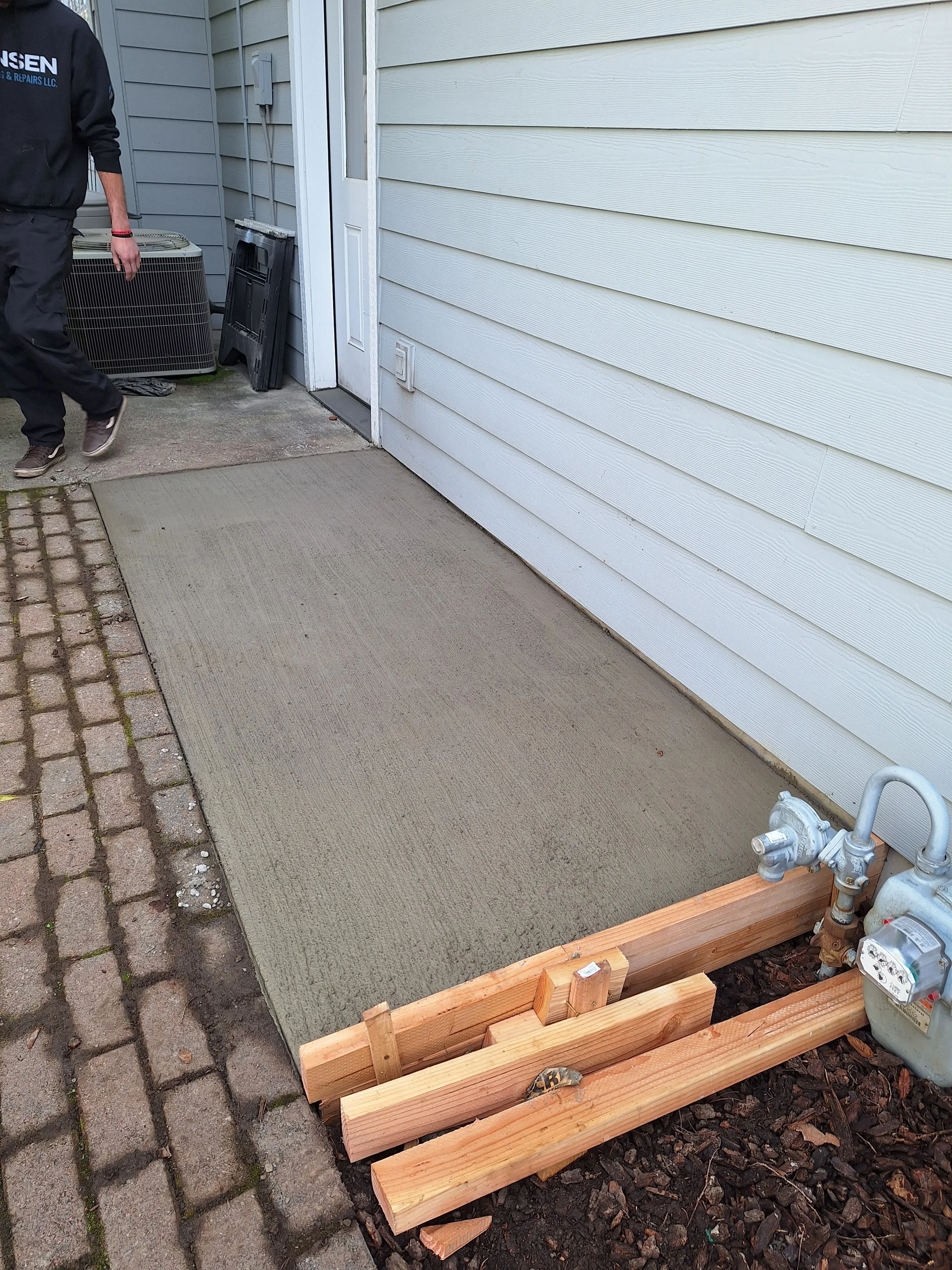 A newly poured concrete patio next to a house with white siding. There is a wooden frame around part of the concrete and an air conditioning unit nearby. A person is partially visible on the left side of the image.