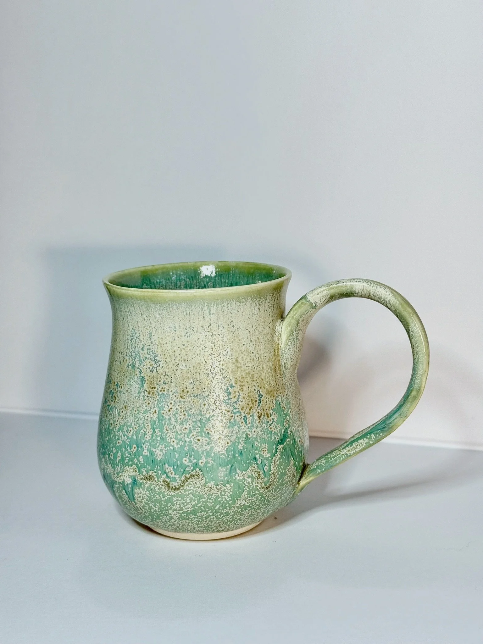 Ceramic pitcher with a textured, green and beige glaze, placed on a white surface against a white background.