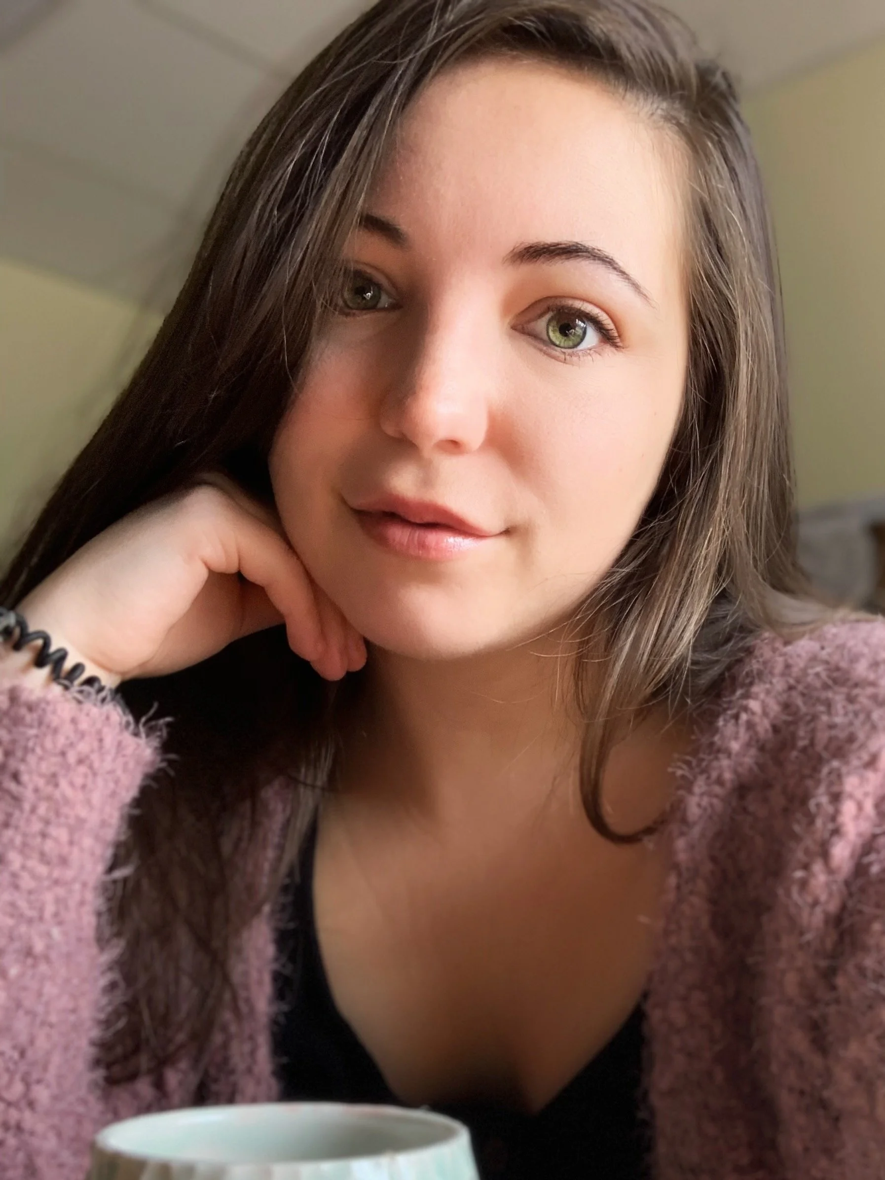 A young woman with long brown hair and green eyes resting her head on her hand, wearing a pink sweater, sitting at a table with a white mug in front of her in a cozy indoor setting.