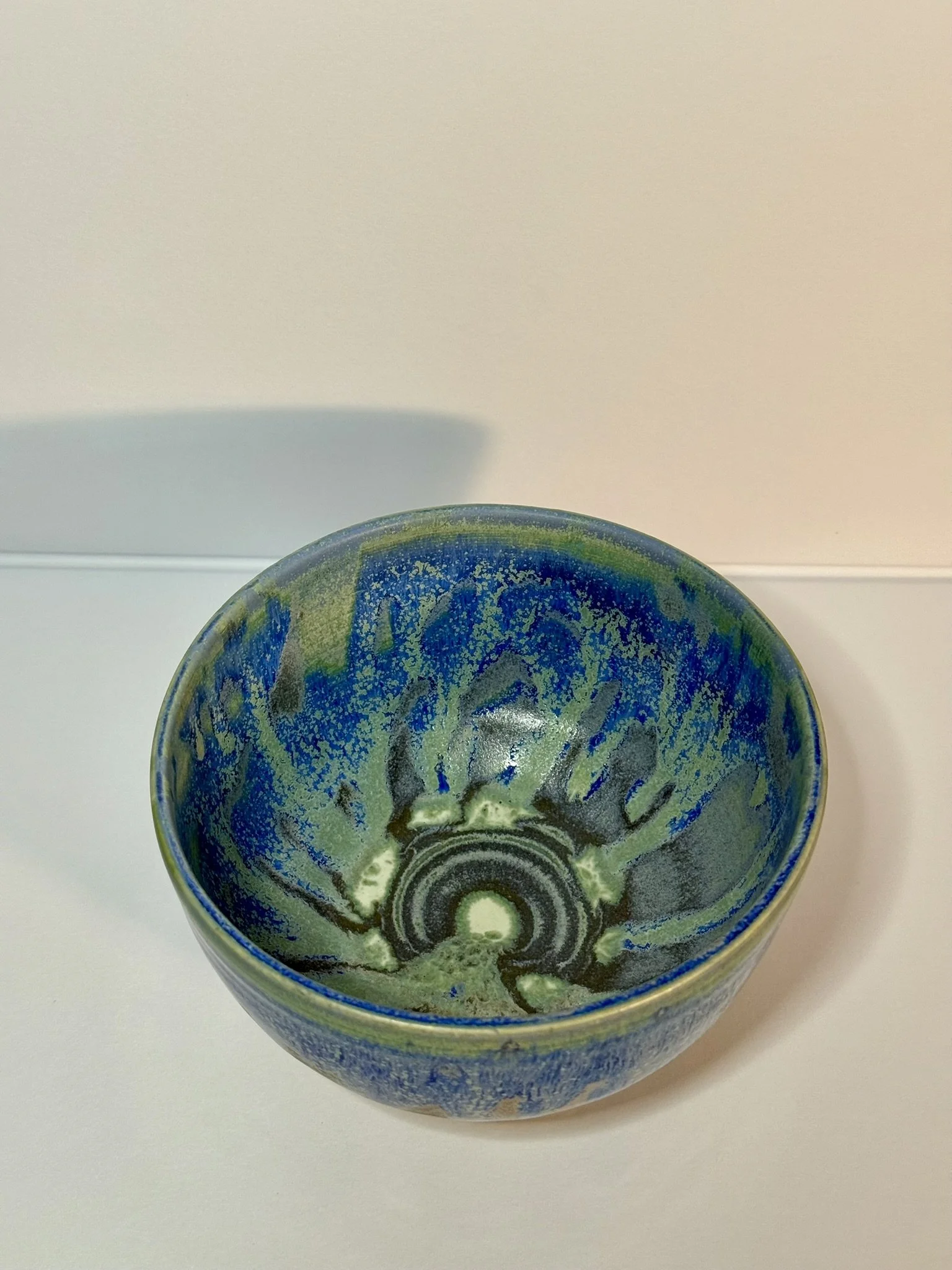 A ceramic bowl with a multicolored glaze pattern in shades of blue, green, and black placed on a white surface against a white background.