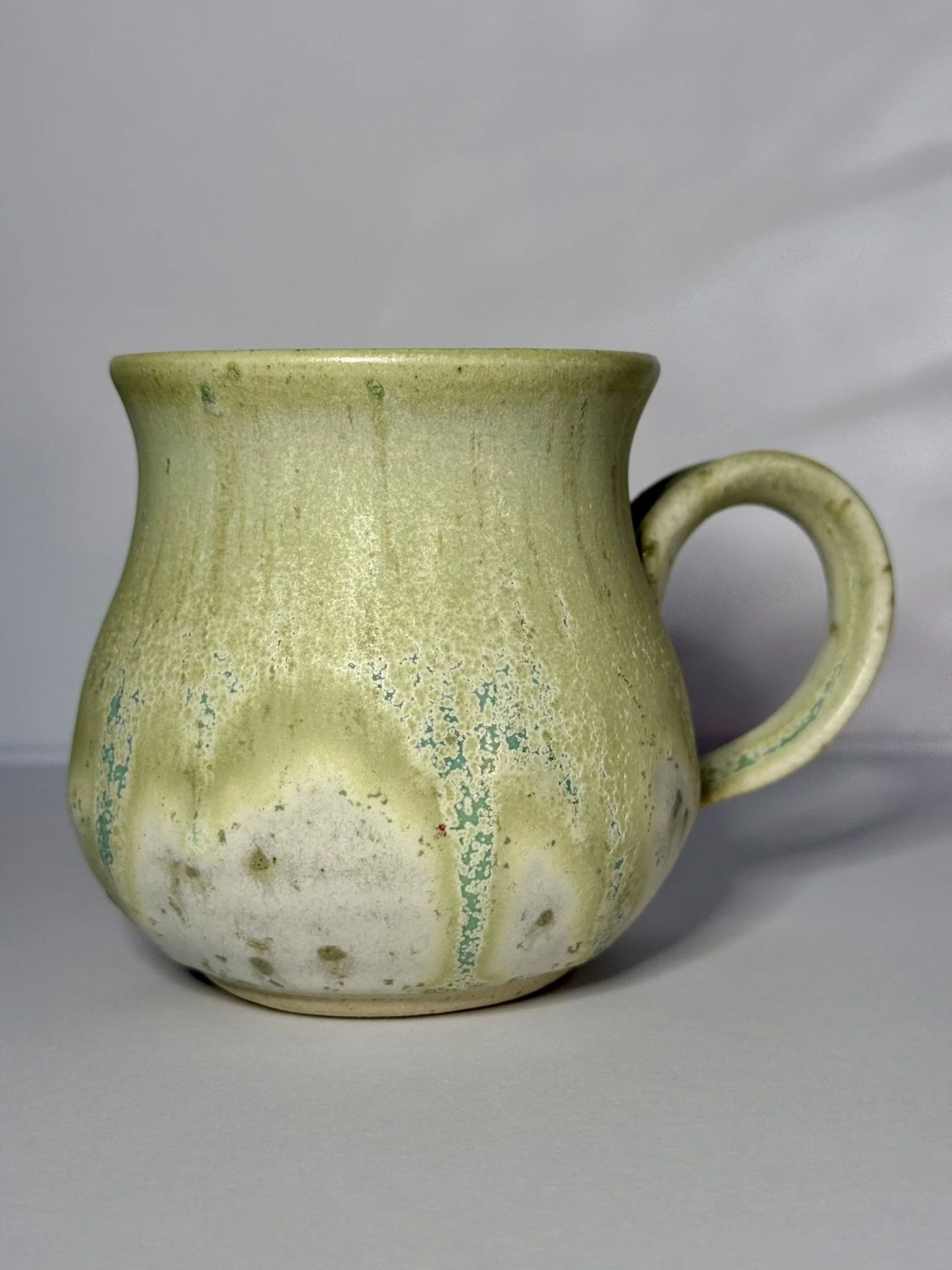 A vintage ceramic mug with a muted green and white glaze, featuring a rounded body and a curved handle, set against a plain gray background.