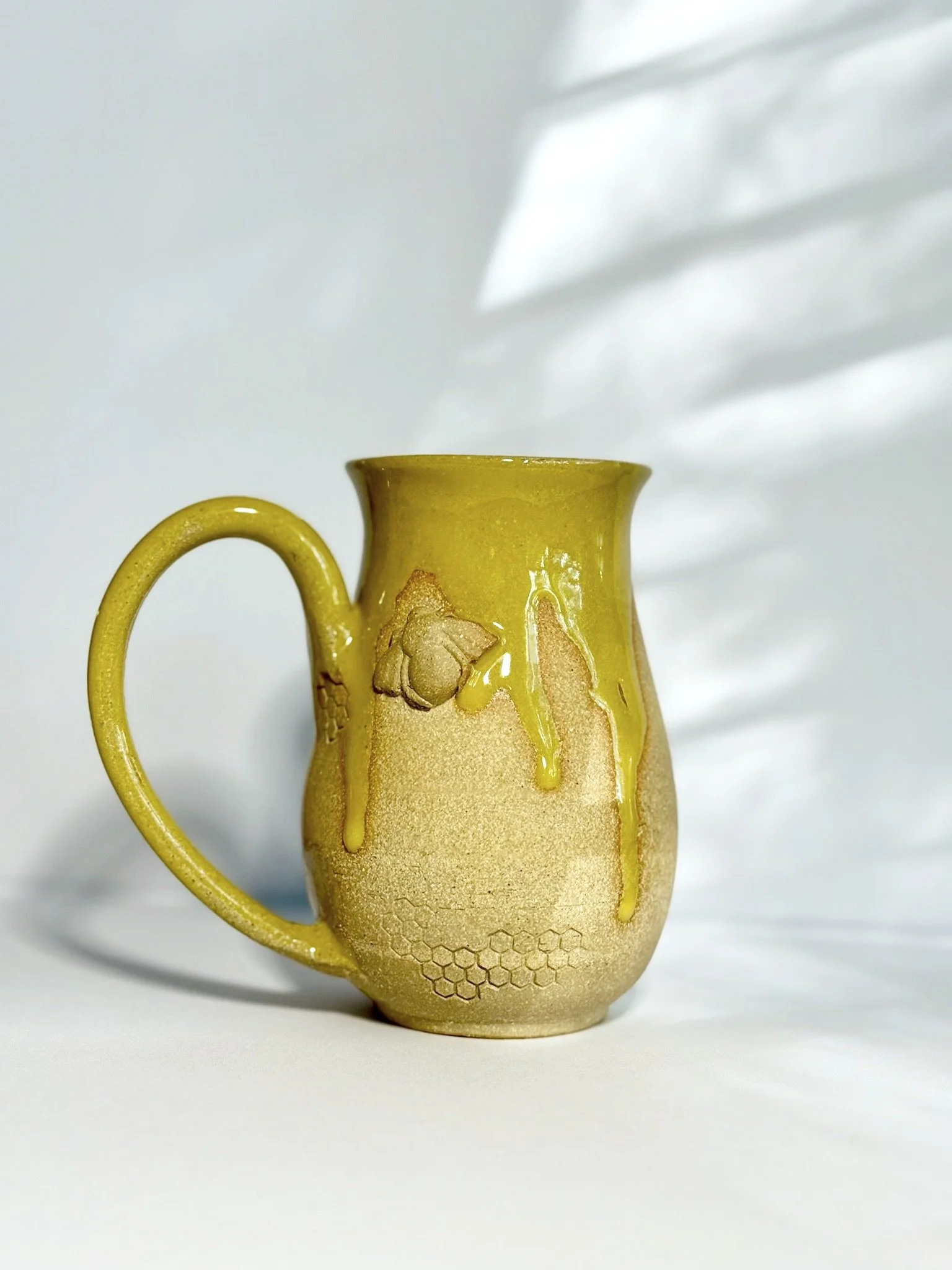Ceramic mug with honeycomb pattern and yellow glaze dripping from the top, placed on a white surface with soft shadows and background.