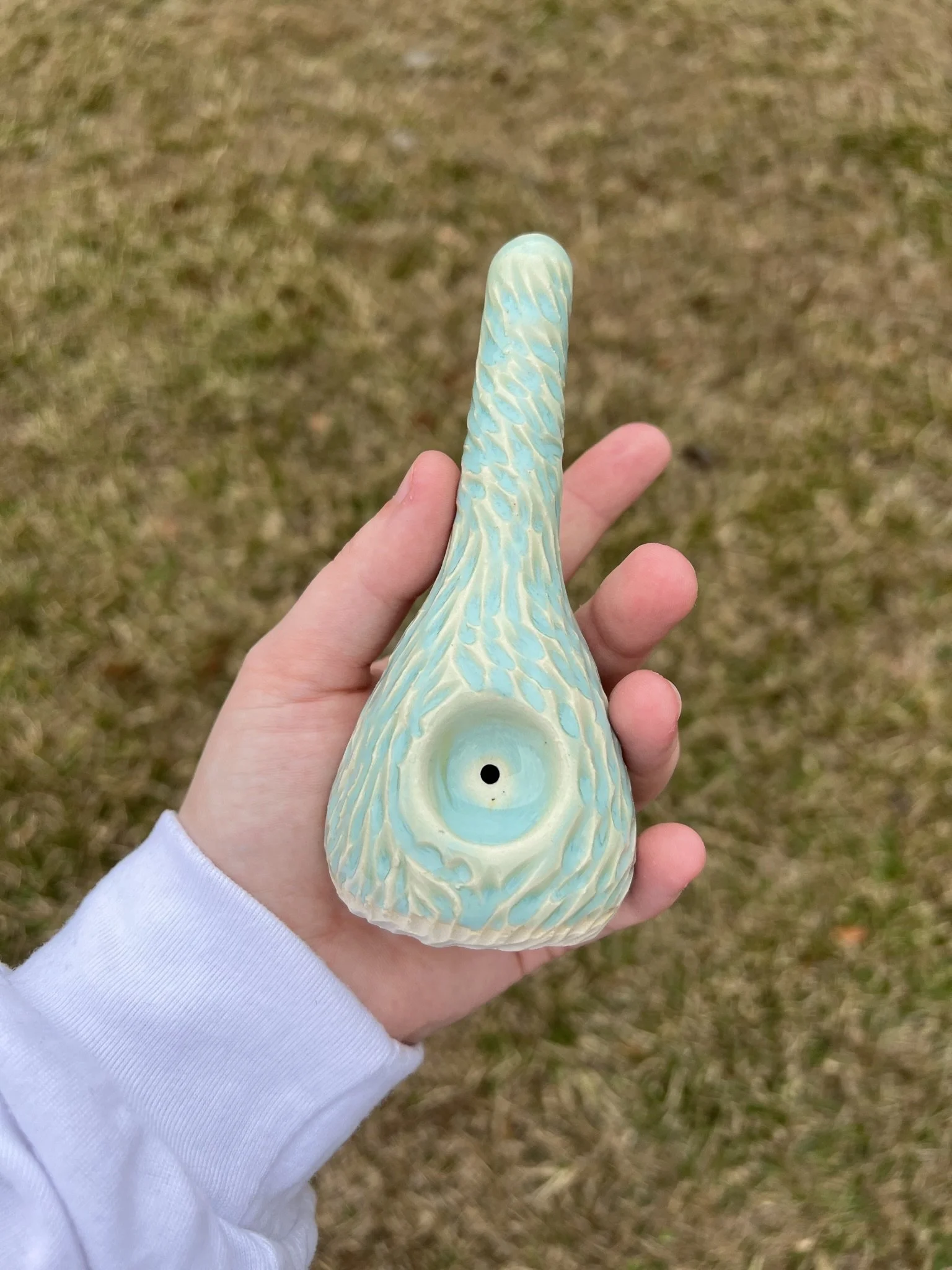Person holding a uniquely designed ceramic smoking pipe with a textured surface, light blue and cream colors, against a grassy background.
