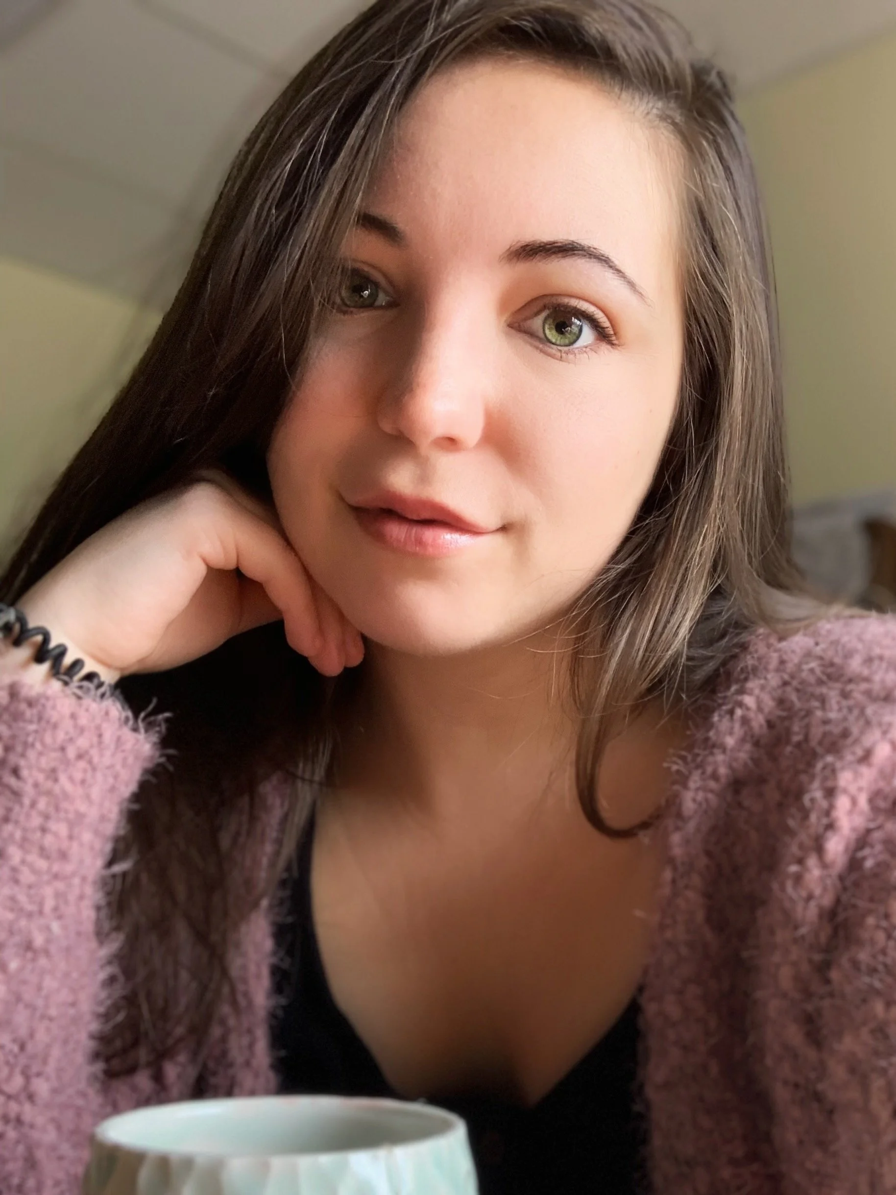 A young woman with long brown hair and green eyes, resting her chin on her left hand, sitting indoors with a mug in front of her.