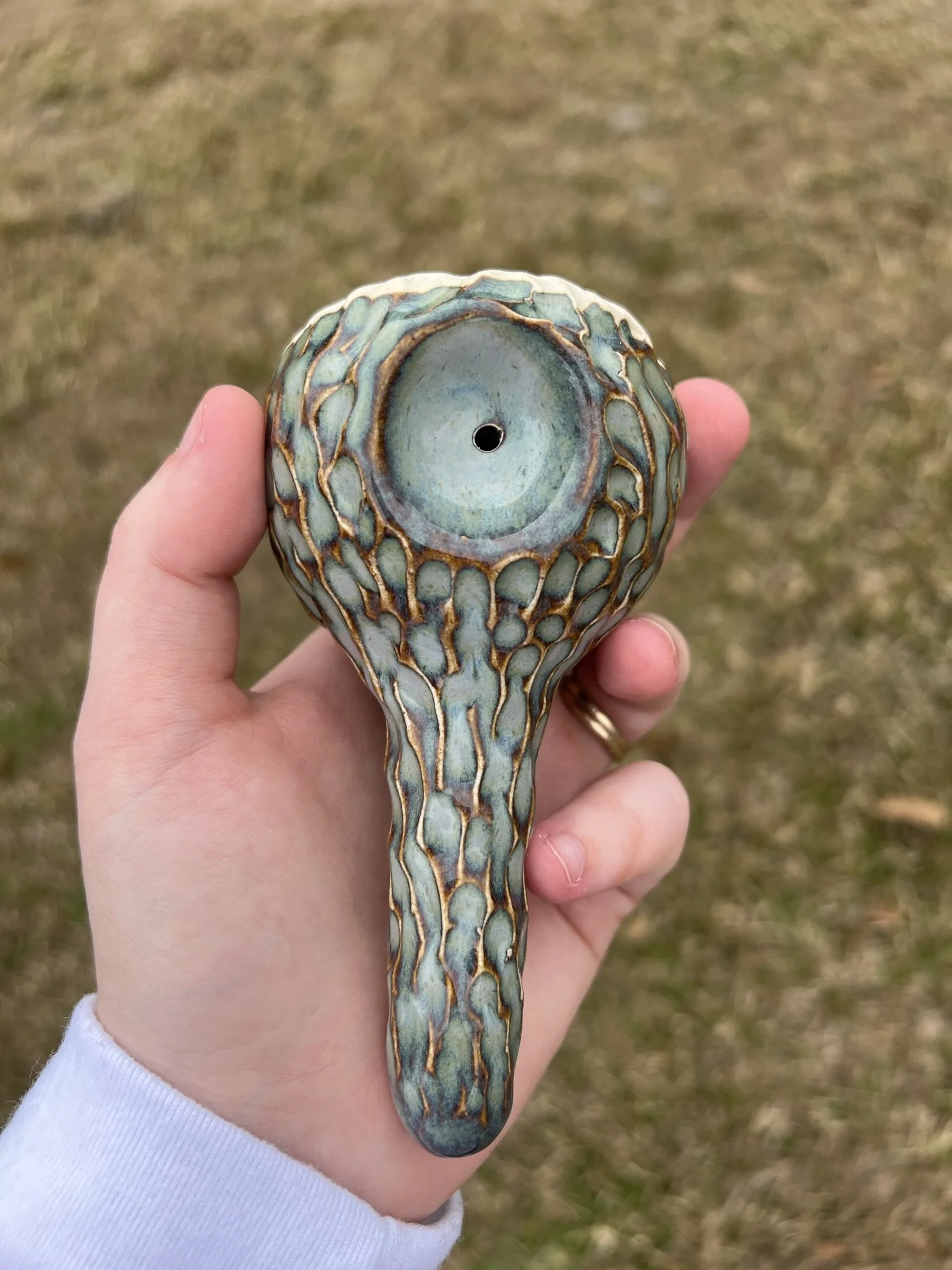A person's hand holding a ceramic water pipe with a textured, reptile skin pattern, outdoors on a grassy surface.