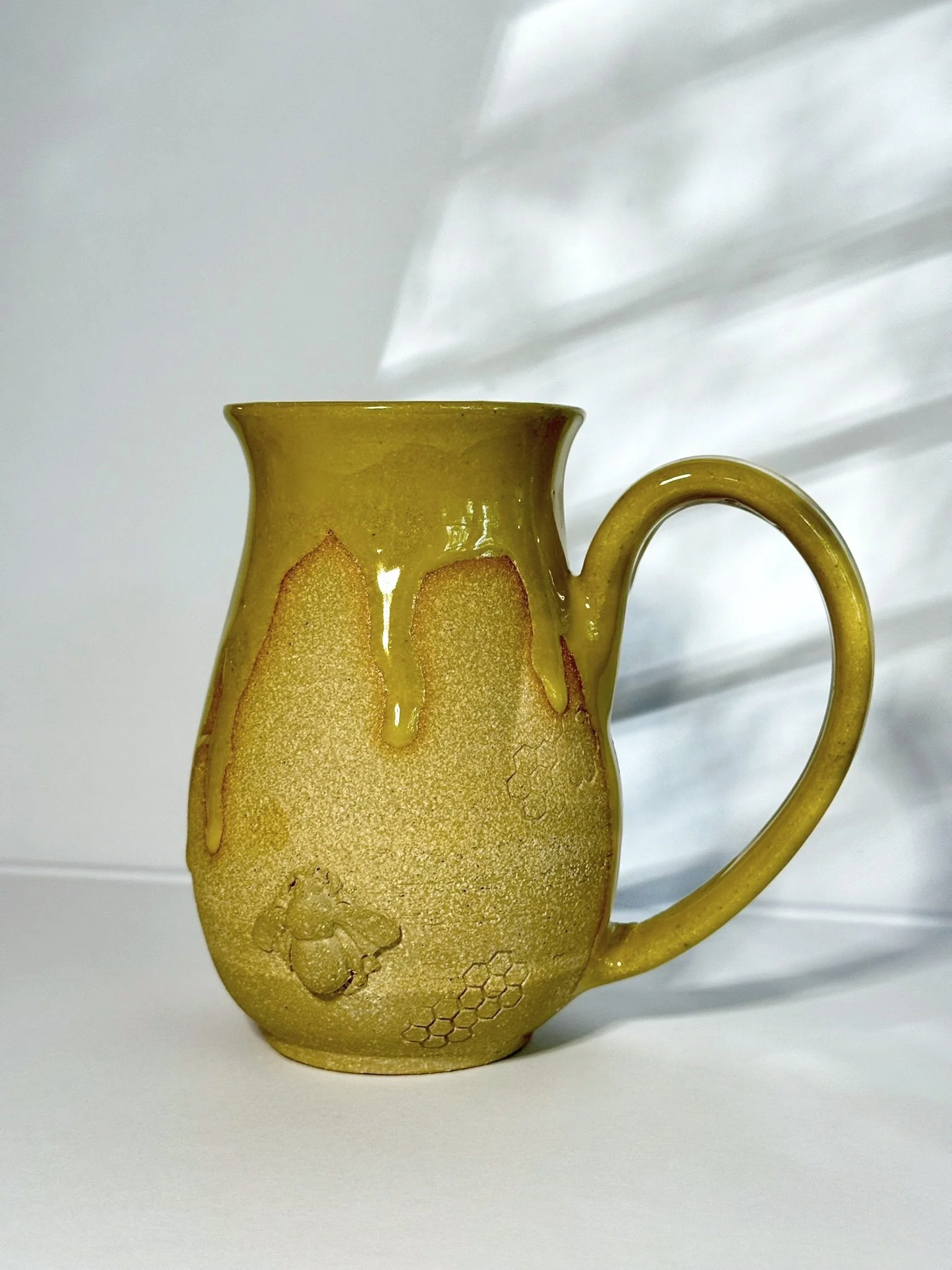 Yellow ceramic mug with honeycomb, bee, and honey dripping design, placed on a white surface with light shadows in the background.