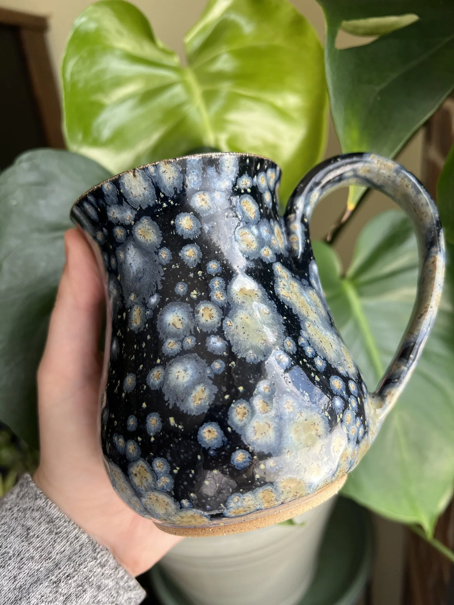Colorful ceramic mug with a black glaze and yellowish and light blue spots, held by a person in front of green leafy plants.