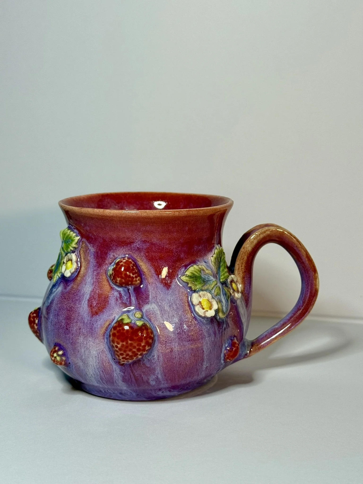 A decorative ceramic mug with a strawberry and floral design, featuring red strawberries, green leaves, and white flowers, set against a plain background.
