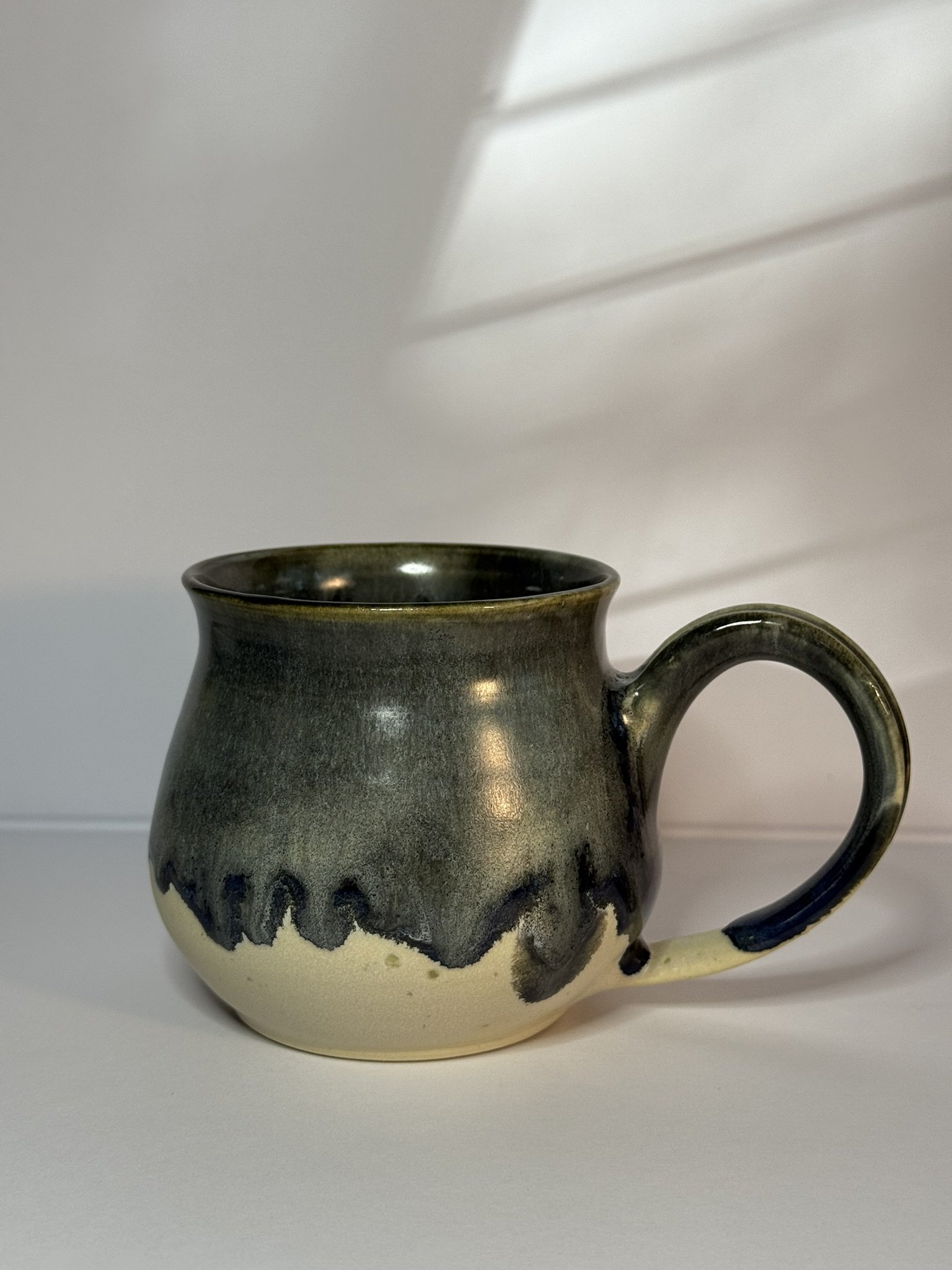 A ceramic mug with a dark green and beige glaze, featuring a wavy pattern along the middle.