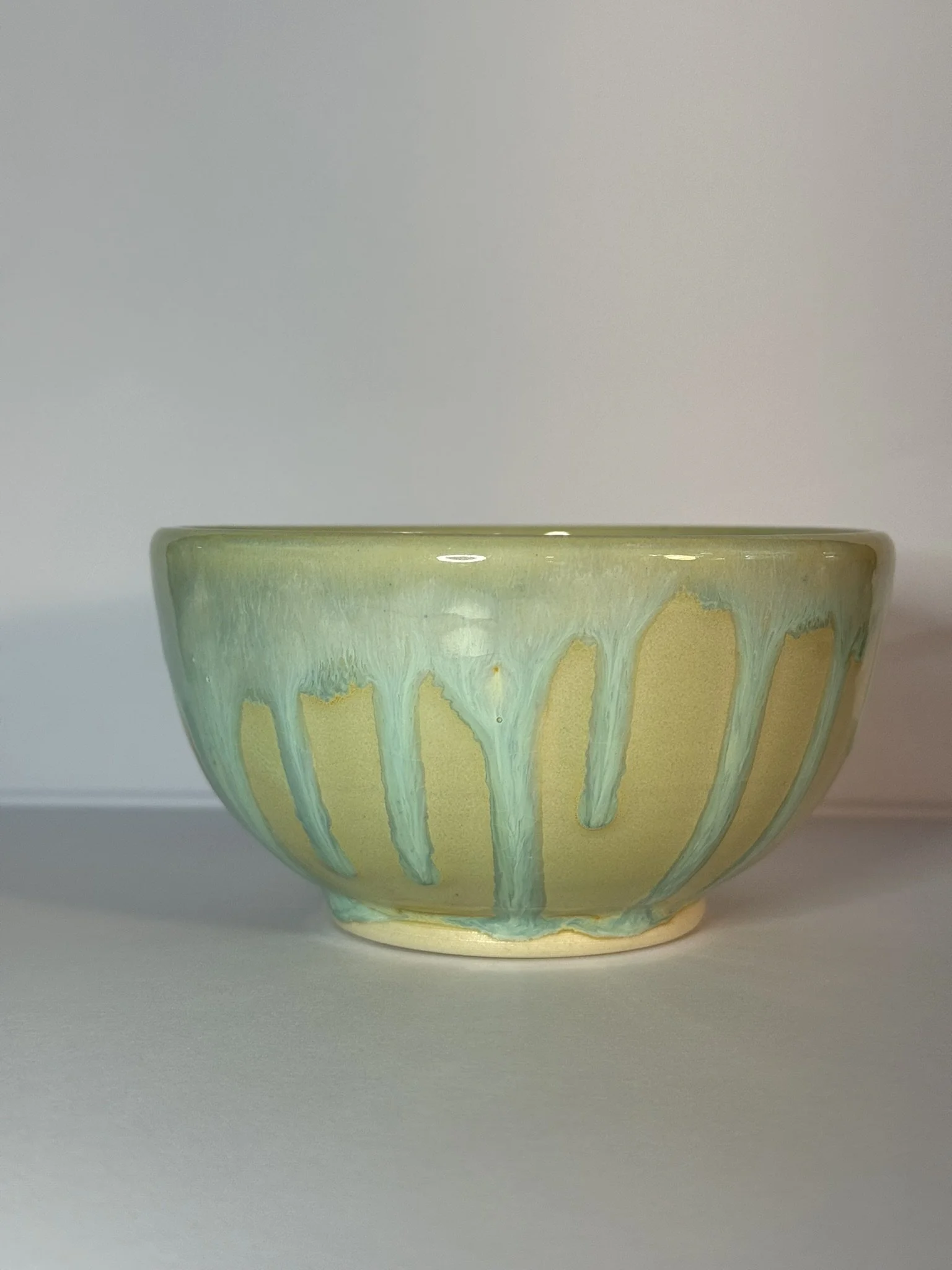A ceramic bowl with a greenish glaze and drippy design on a white surface against a plain background.