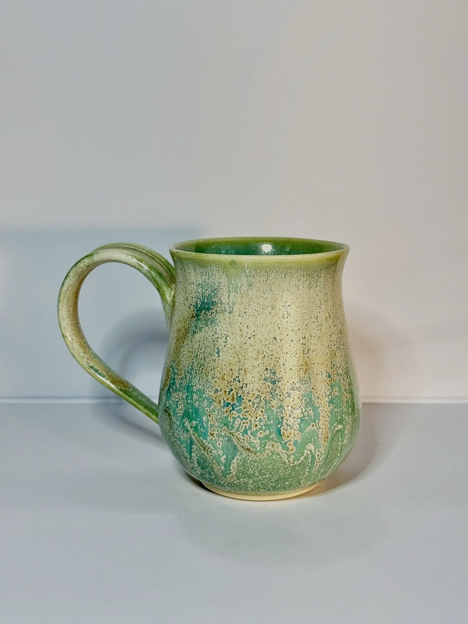 Green and cream ceramic mug with a textured, speckled pattern, placed on a white surface against a neutral background.