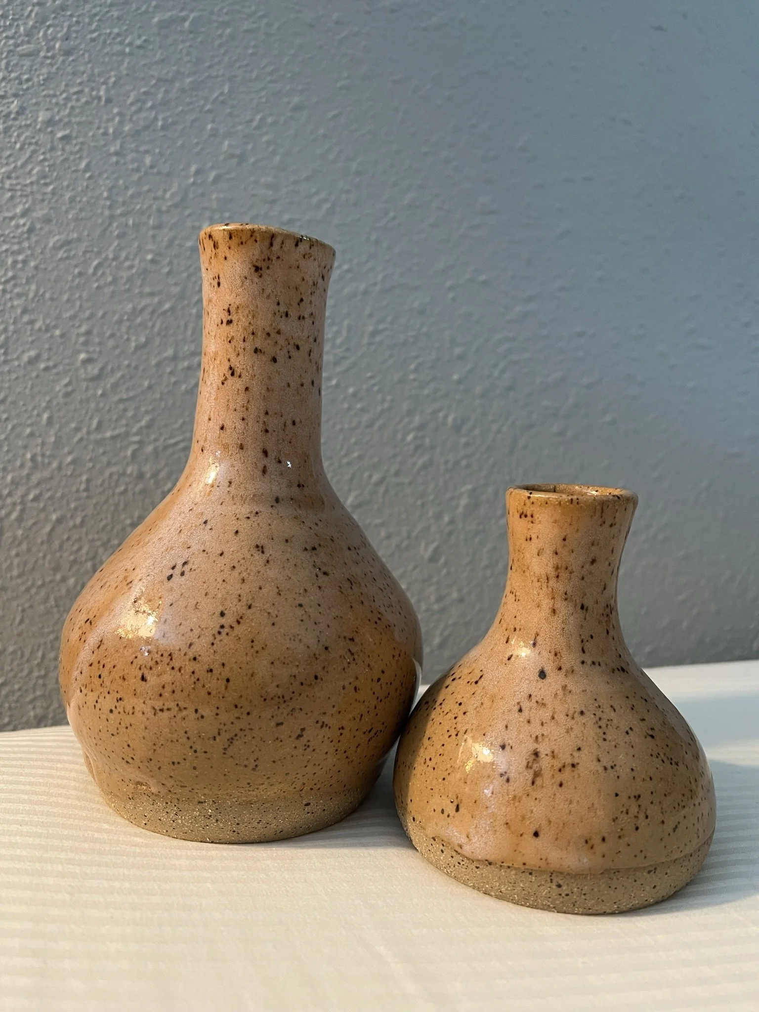 Two beige speckled ceramic vases on a white surface against a gray wall.