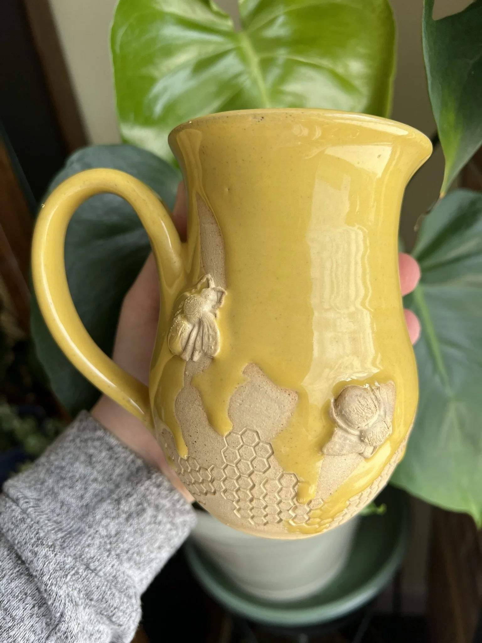 Yellow ceramic mug with bee and honeycomb design, being held by a person wearing a gray sleeve, with green leafy plants in the background.