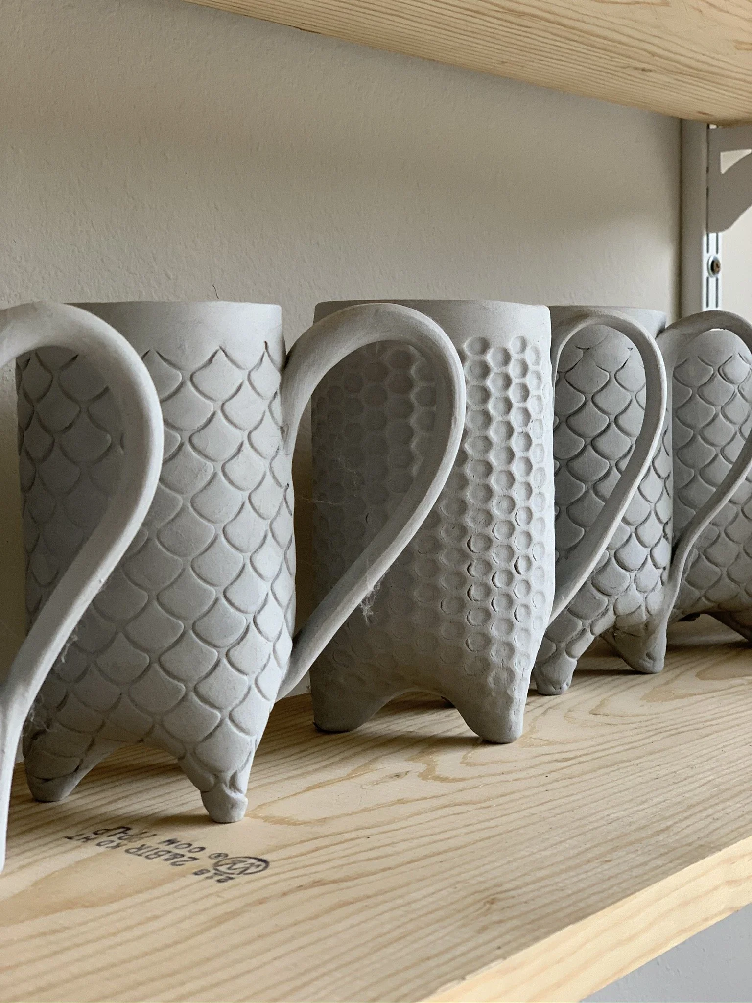 Multiple white ceramic mugs with textured honeycomb and scale patterns on a wooden shelf.