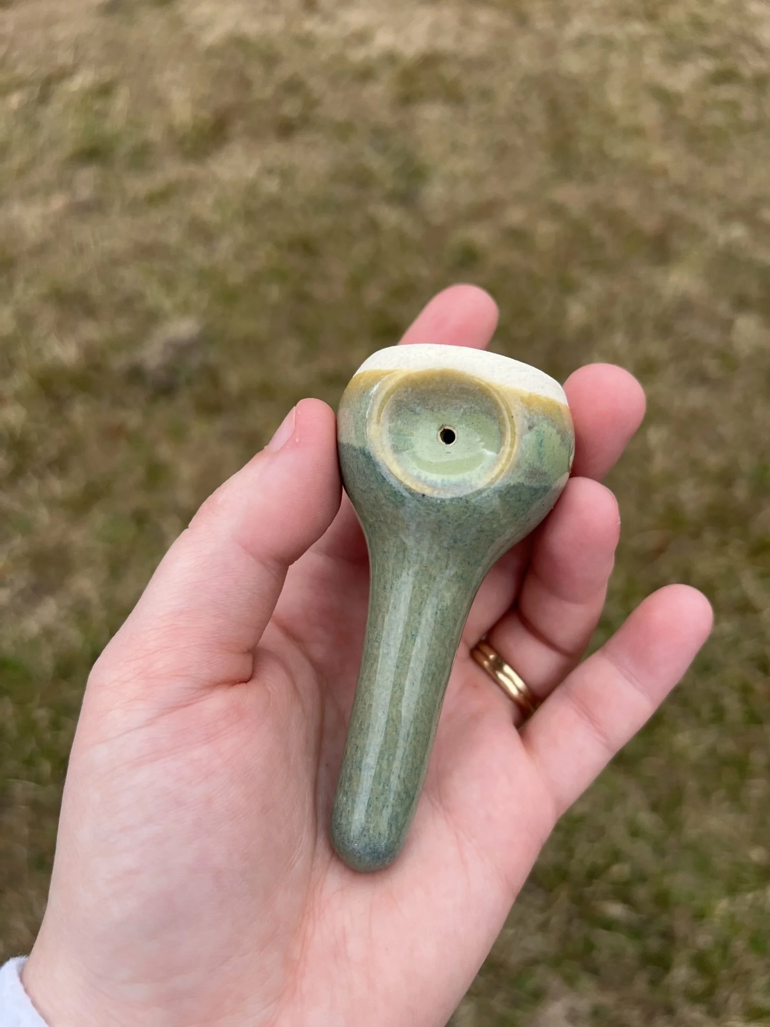 Hand holding a small ceramic object with a circular hole and a textured, greenish surface, outdoors on a grassy background.