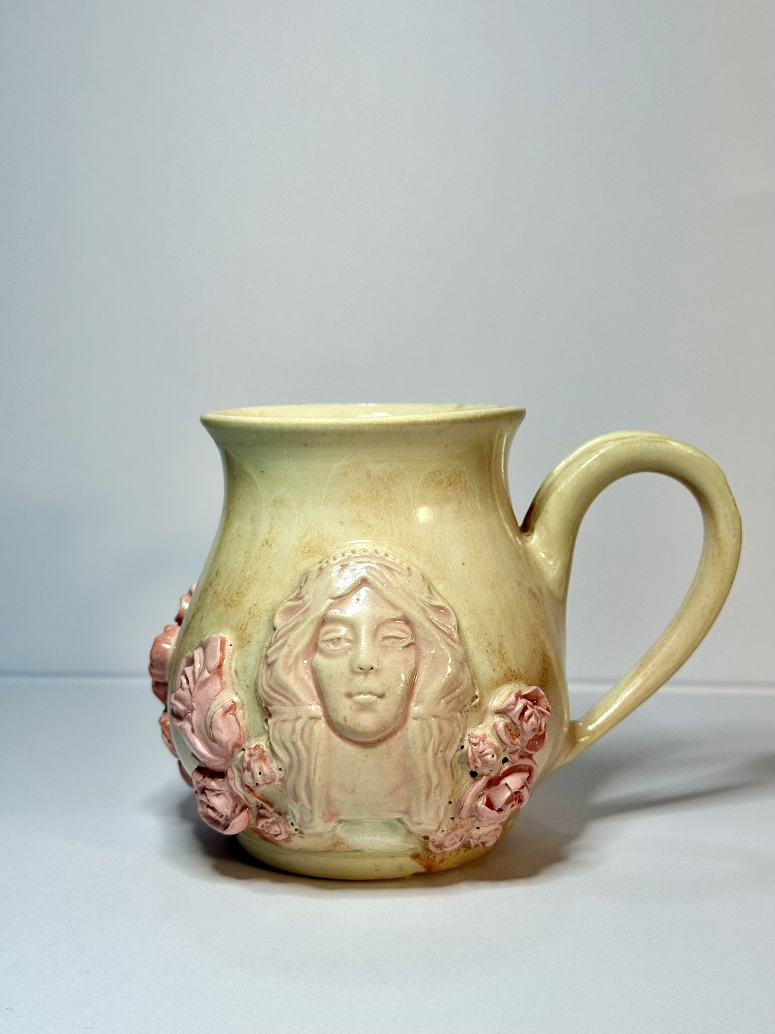 A ceramic mug featuring a relief of a woman's face with wavy hair, surrounded by pink roses, on a plain background.
