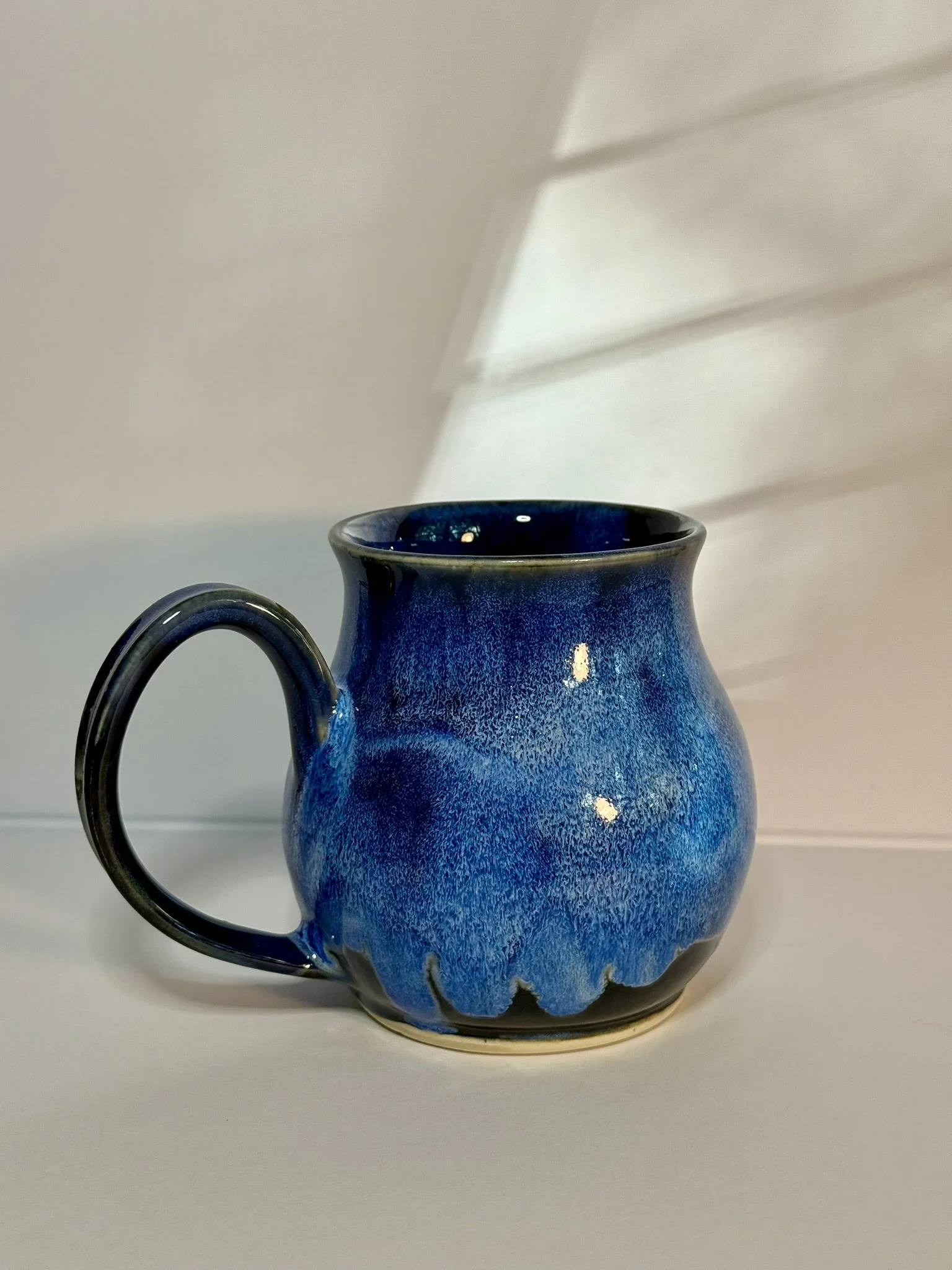 Blue ceramic mug with black handle on a white surface, with a white wall and window blinds in the background.