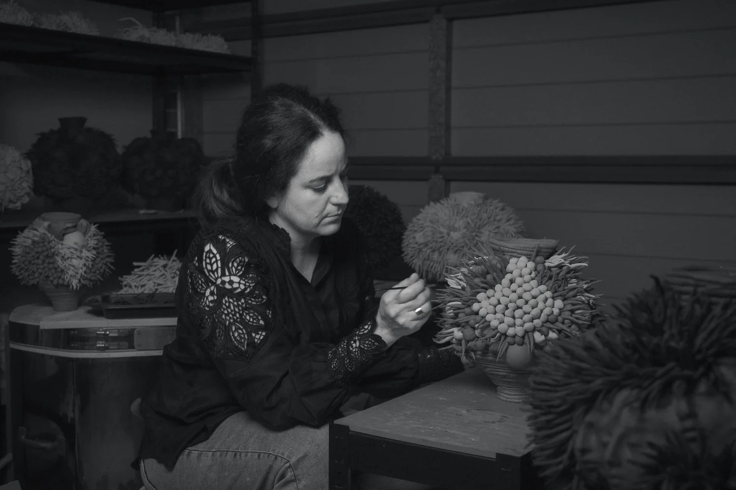 A woman artist with dark hair working on a ceramic floral arrangement in a studio surrounded by various spherical artworks.