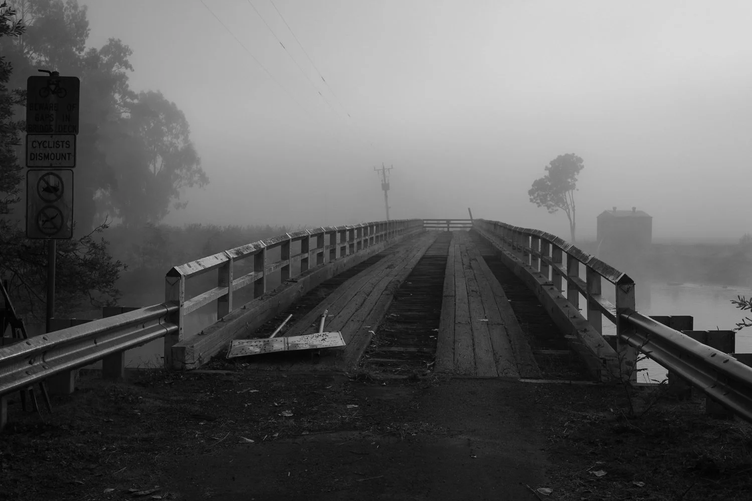 A foggy scene featuring a small wooden bridge over a body of water, with attached signs on the left, a house in the distance, and trees shrouded in fog.