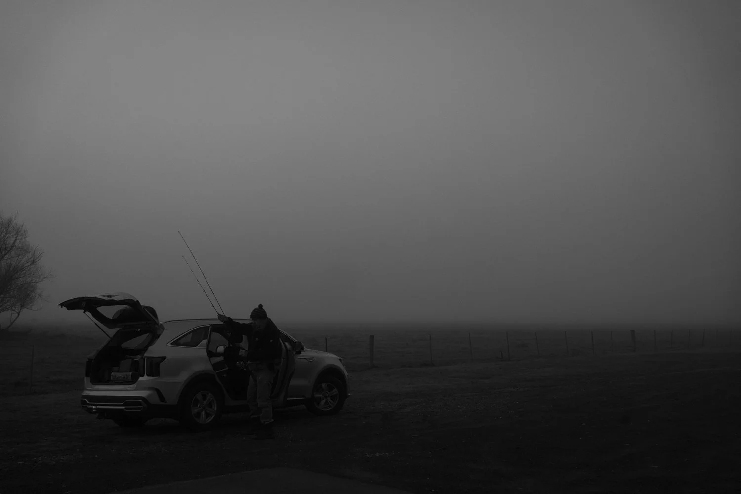 A person opening a car trunk in a foggy outdoor setting, with trees and a fence in the background.