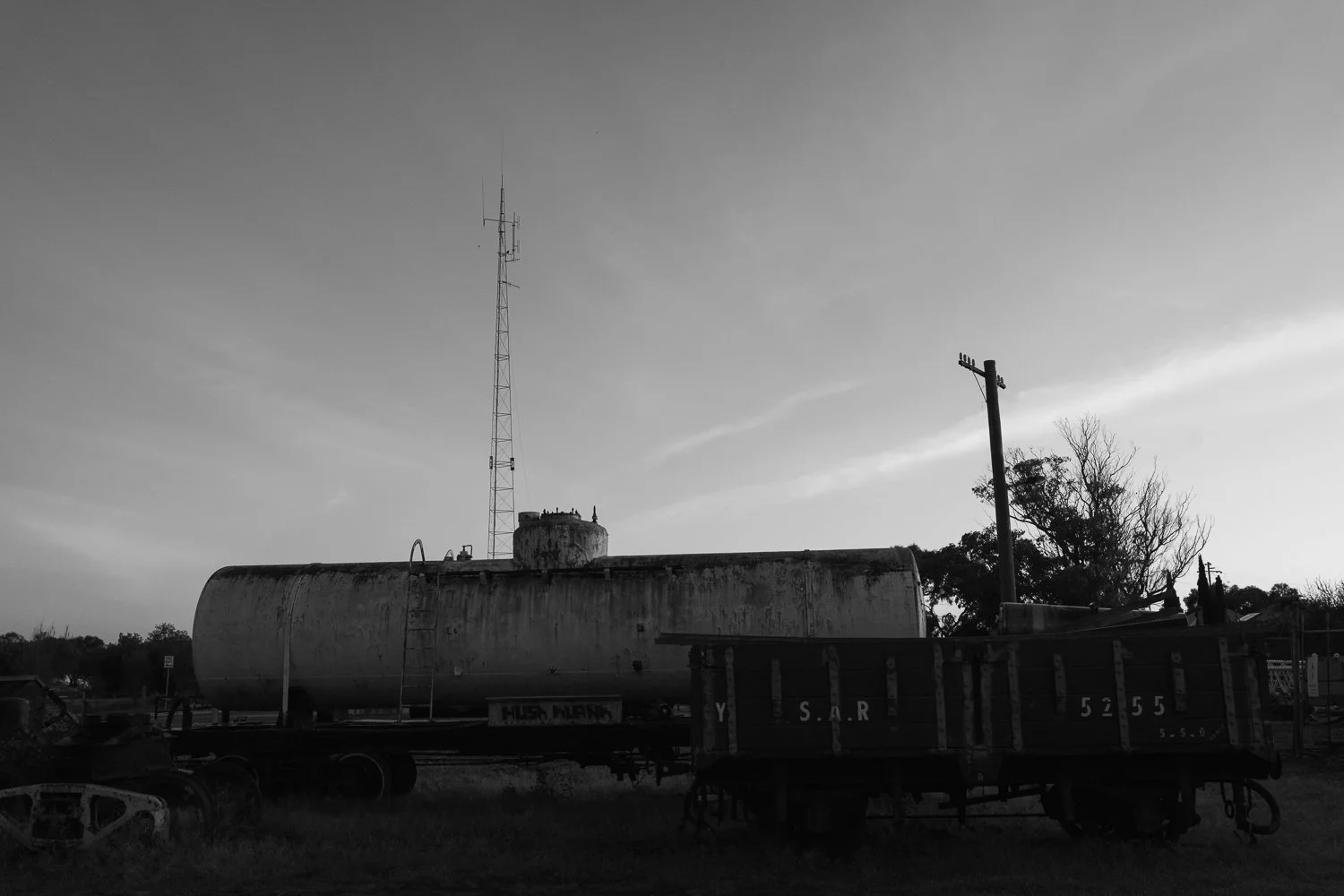 Black and white photo of an old train car and tanker on railroad tracks, with a communication tower and a utility pole in the background, and trees in the distance.