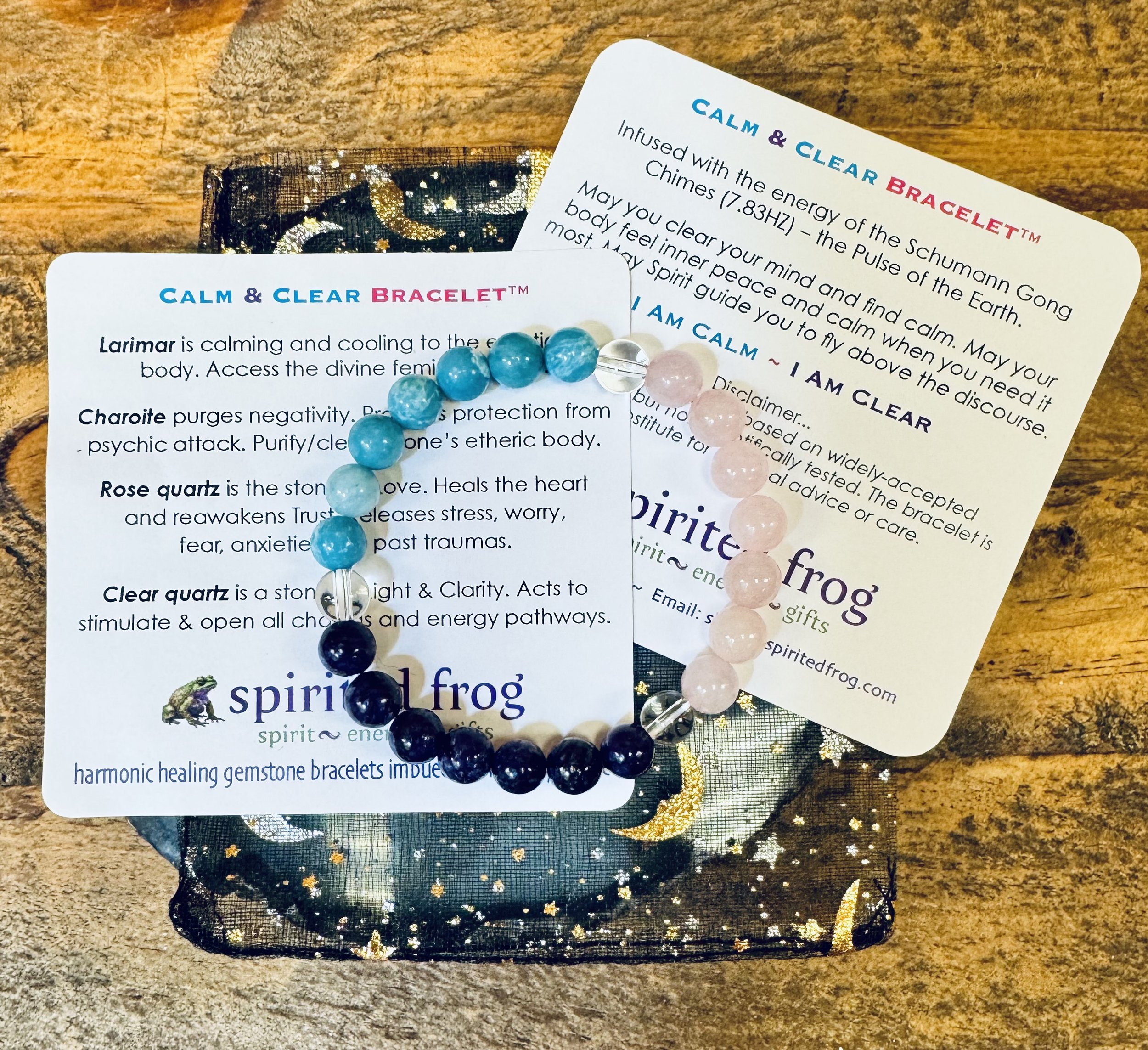 A gemstone bracelet in blue, pink and purple are shown on top of informational cards about healing crystals; a mesh pouch with star patterns is partially visible behind the cards, on a wooden surface.
