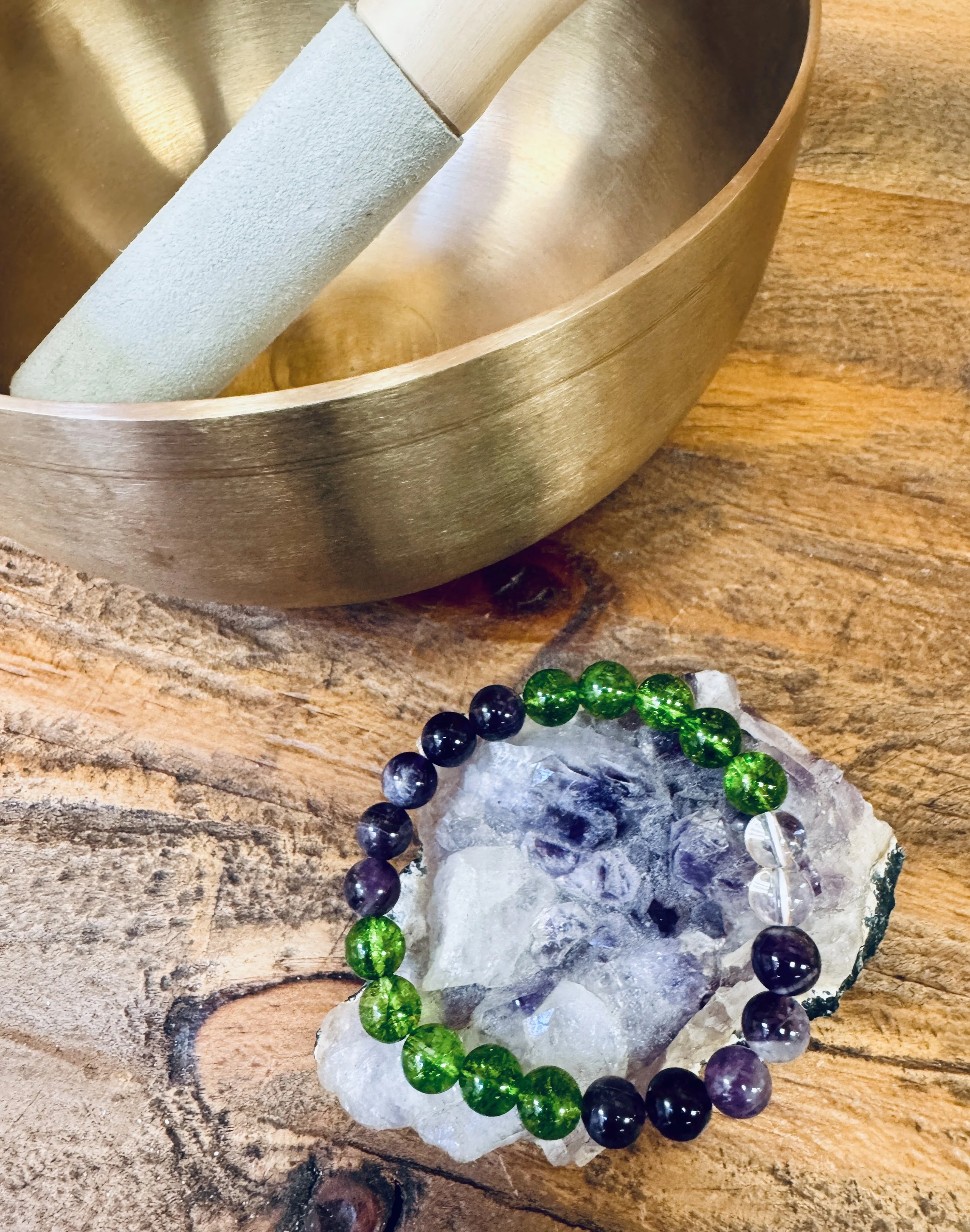 A wooden table with a brass bowl, a large clear rock, and a beaded bracelet with green and black beads.