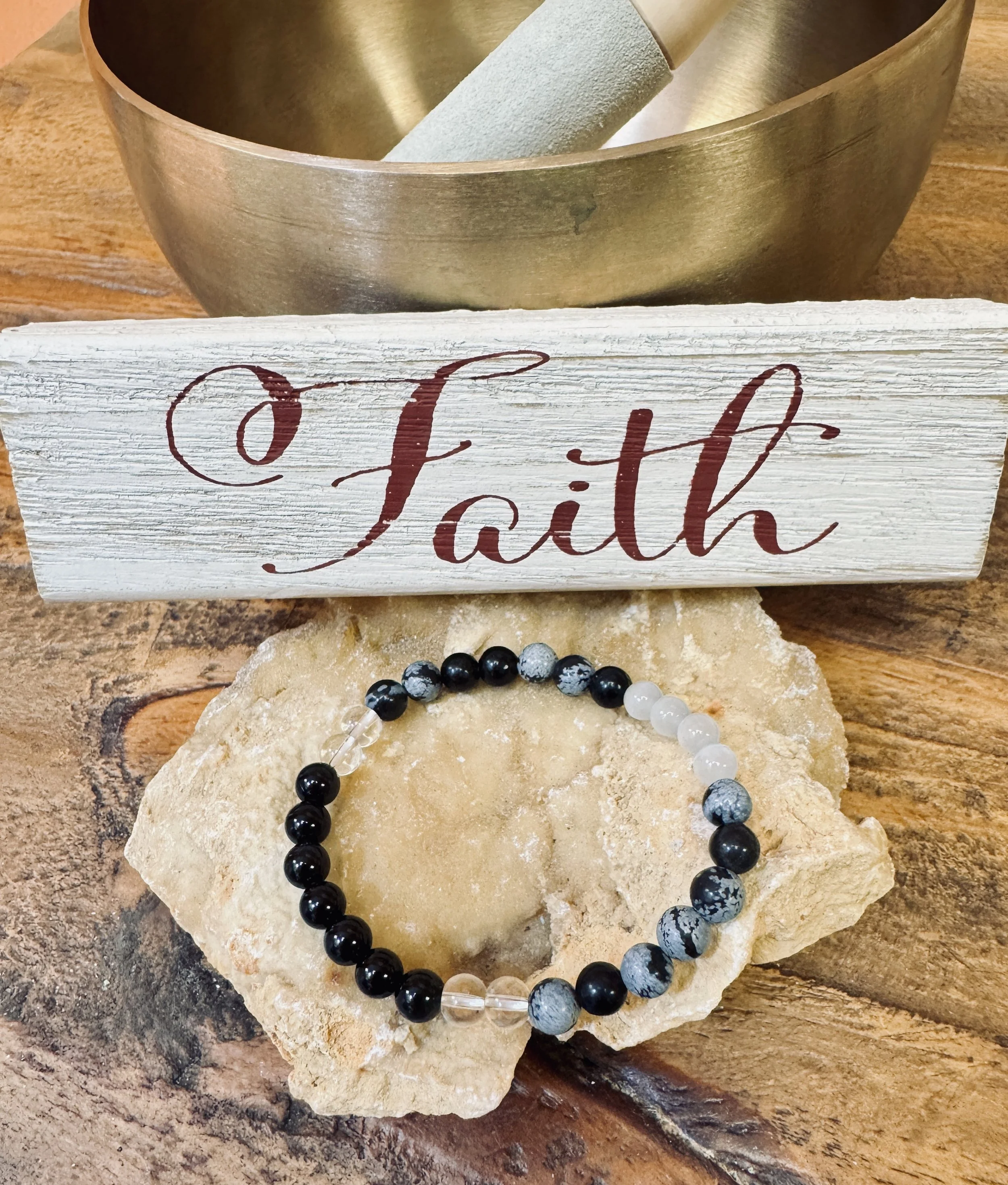 A black and gray beaded bracelet resting on a large beige rock, with a wooden and white painted decorative sign that reads 'Faith' placed behind it.